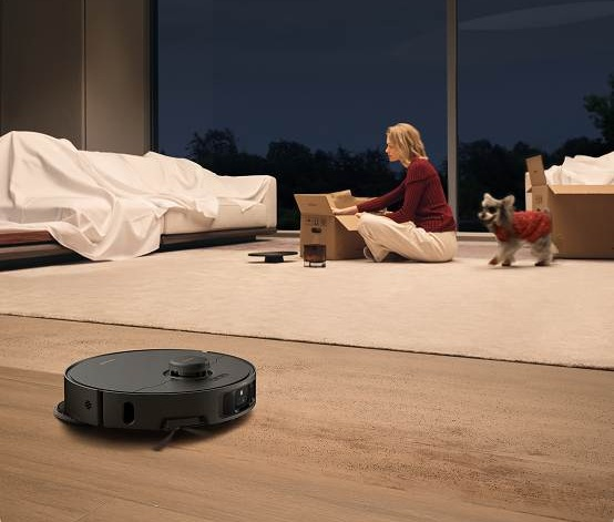 Kobieta rozpakowuje karton, mały pies chodzi, a robot sprzątający stoi na podłodze. W tle kanapa.