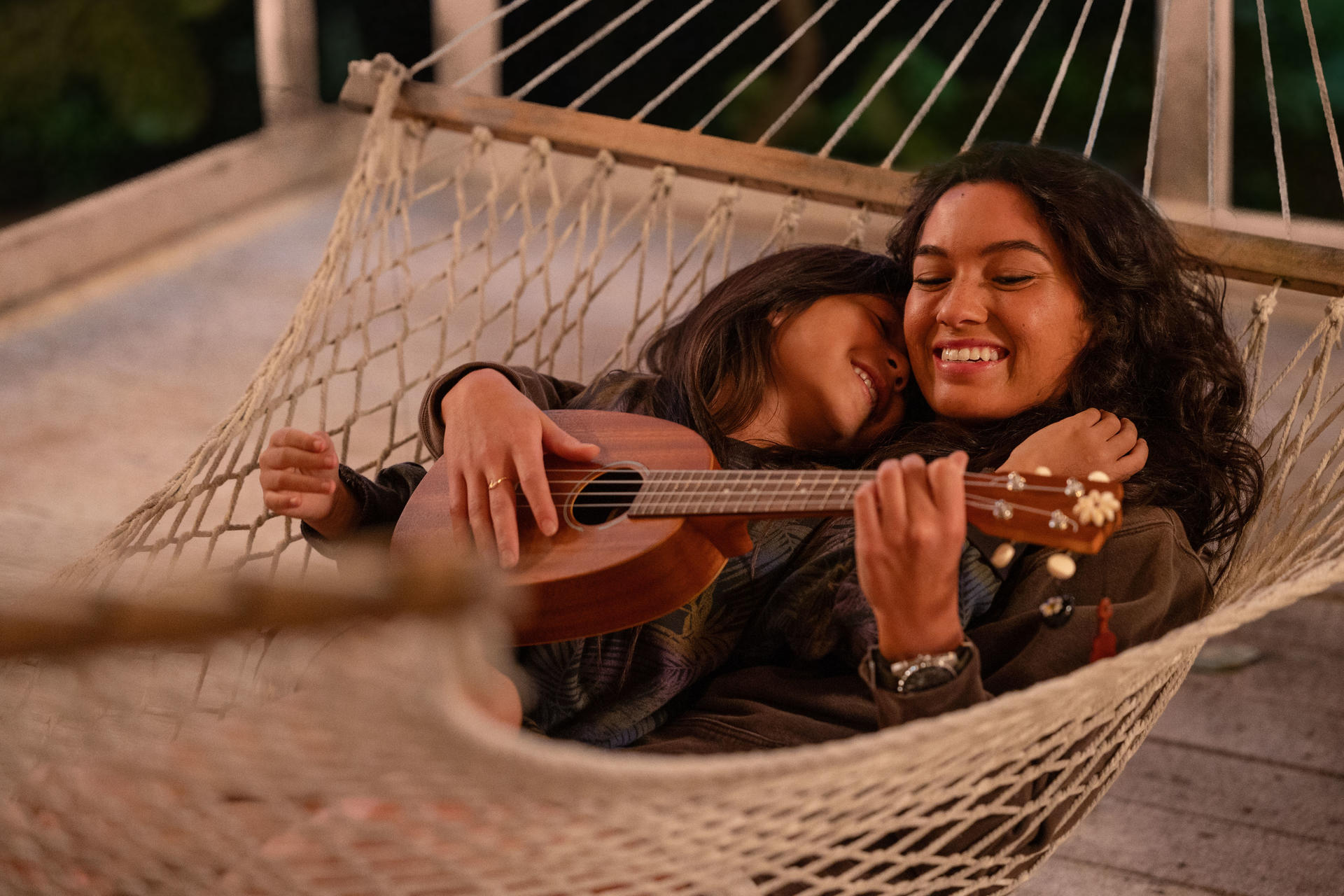 Zwei Frauen in einer Hängematte, eine spielt Ukulele. Sie lächeln und umarmen sich. Die Szene wird von weichem, natürlichem Licht erhellt.