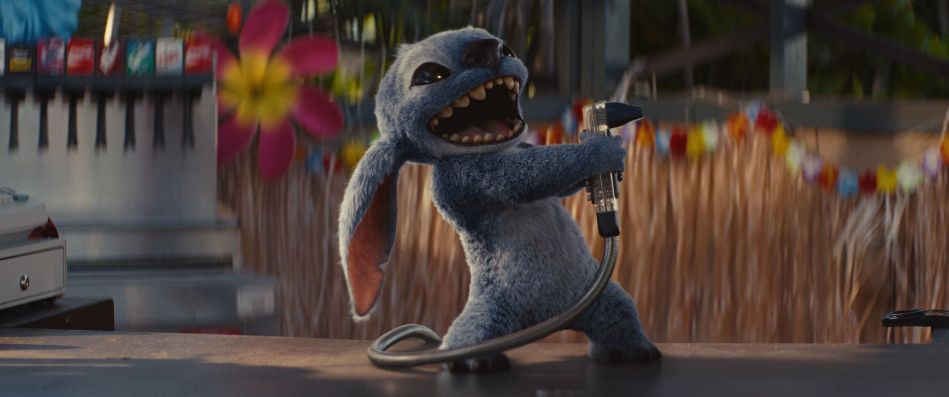 Stitch hält einen Getränkespender. Im Hintergrund befinden sich farbenfrohe Girlanden- und Blumendekorationen. Der Tresen ist im Vordergrund.