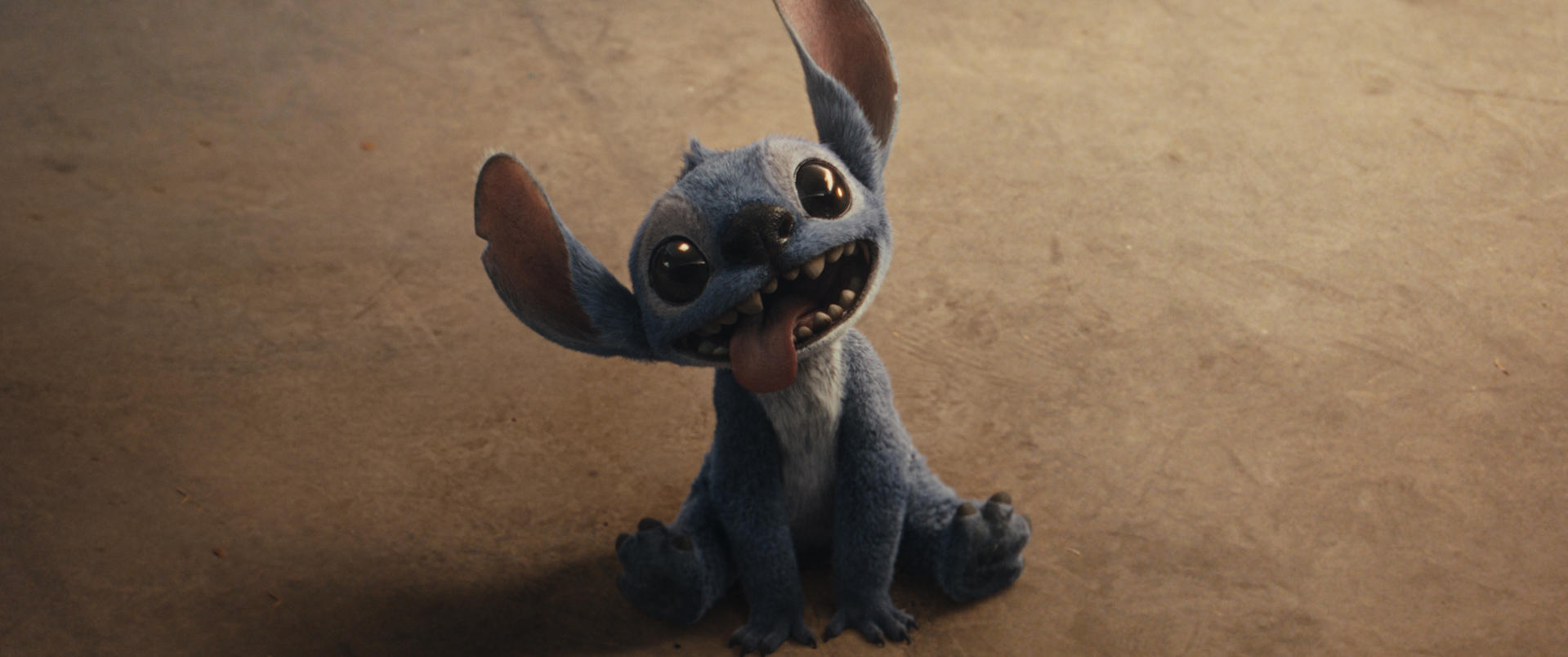 Stitch, blauer Außerirdischer, lächelt mit herausgestreckter Zunge. Er sitzt auf einer braunen Oberfläche. Große Augen und Ohren. Der Hintergrund ist unscharf.