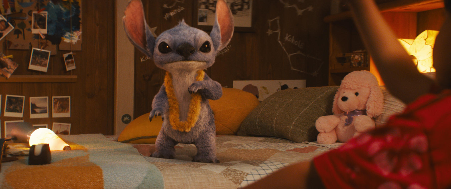 Stitch steht auf einem Bett und trägt eine Blumengirlande. Ein rosa Pudel-Plüschtier sitzt daneben. Fotos hängen an der Holzwand.