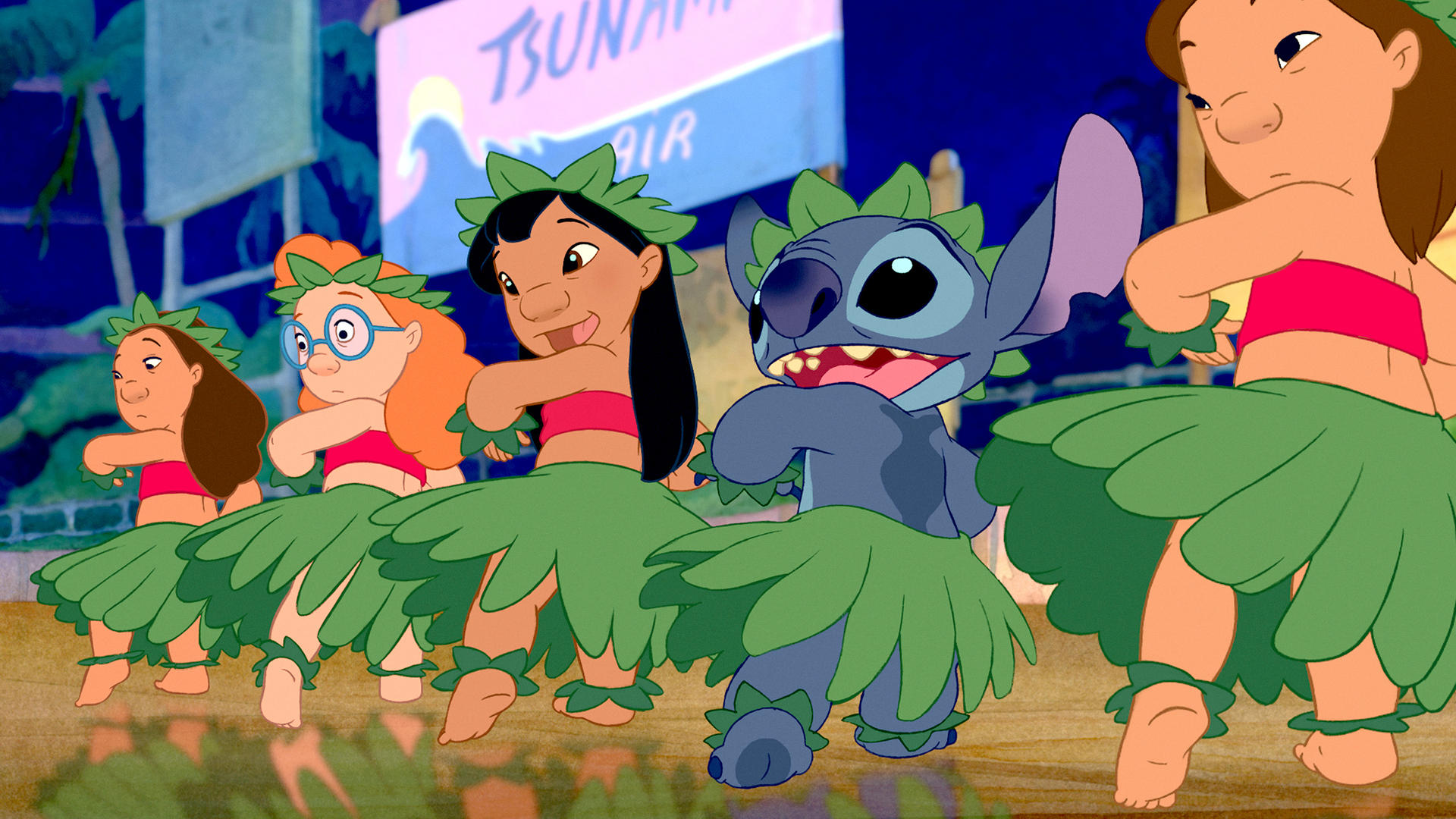 Eine Gruppe von Kindern und Stitch tanzen in traditioneller Kleidung. Sie tragen Grassröcke, Blattkränze und rote Oberteile. Im Hintergrund ist ein Schild zu sehen.