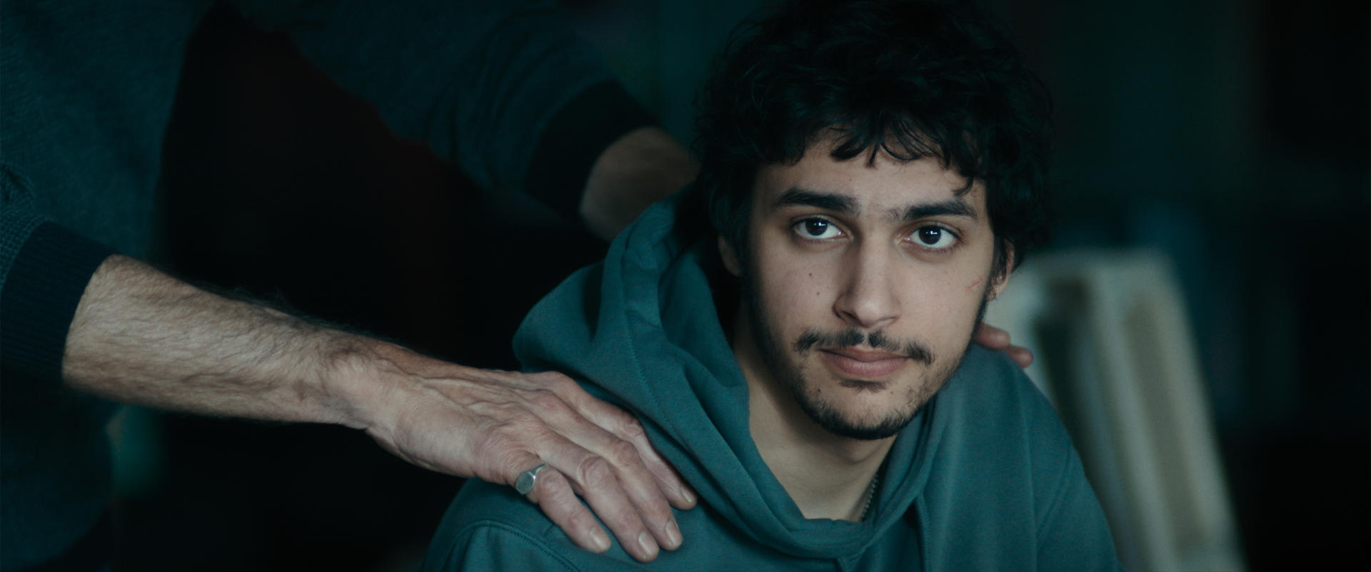 Ein junger Mann in einem teal Hoodie wird von einer Hand auf seiner Schulter gestützt. Die Szene ist schwach beleuchtet, was eine düstere Stimmung erzeugt.