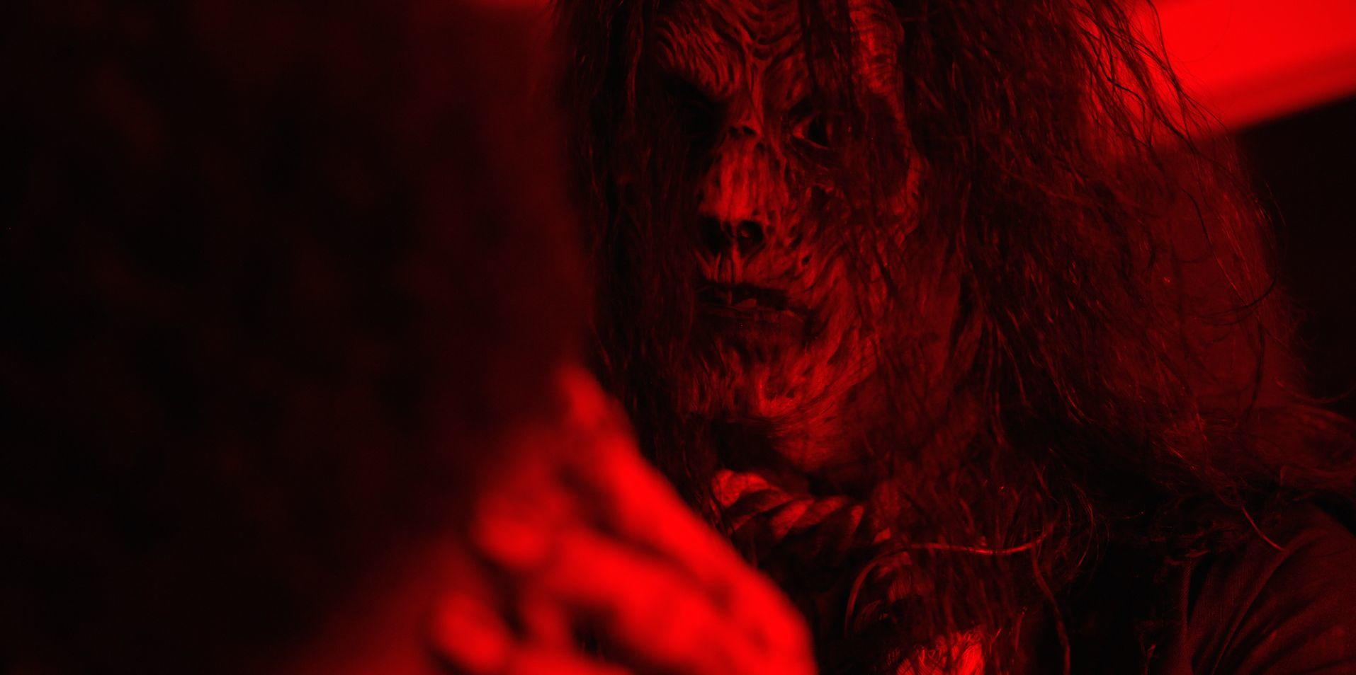 Ein bleiches Wesen mit langen, dunklen Haaren blickt nach vorn. Rotes Licht durchflutet die Szene. Eine Hand ist im Vordergrund sichtbar.