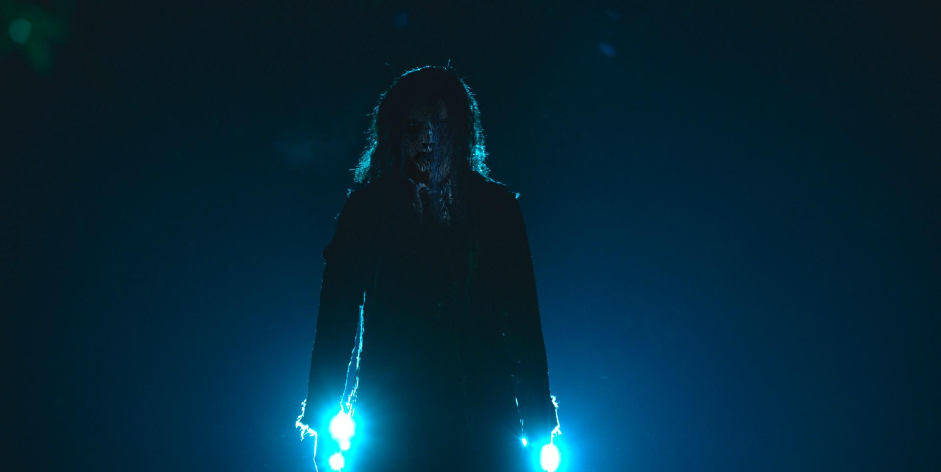 Eine silhouettehafte Figur mit langen Haaren und blassem Gesicht steht vor einem dunkelblauen Hintergrund, der von hellblauen Lichtern beleuchtet wird.