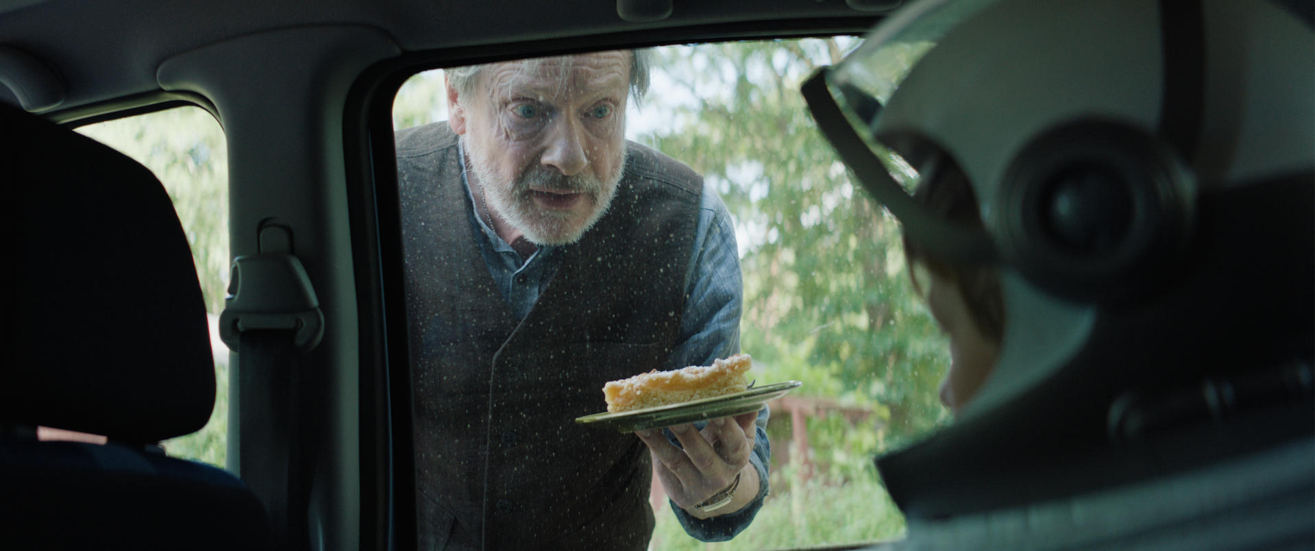 Ein älterer Mann im Autofenster bietet jemandem mit Helm Kuchen an. Der Hintergrund zeigt Bäume durch das Fenster.