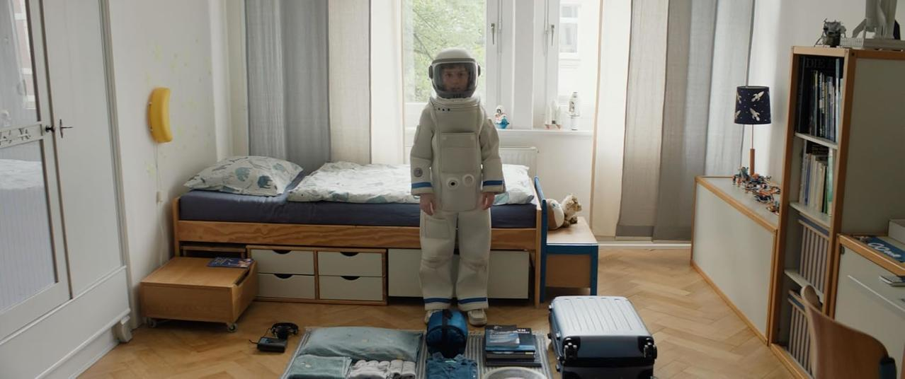 Kind in Raumanzug steht neben Bett mit Gepäck, Zimmer mit Bücherregal.