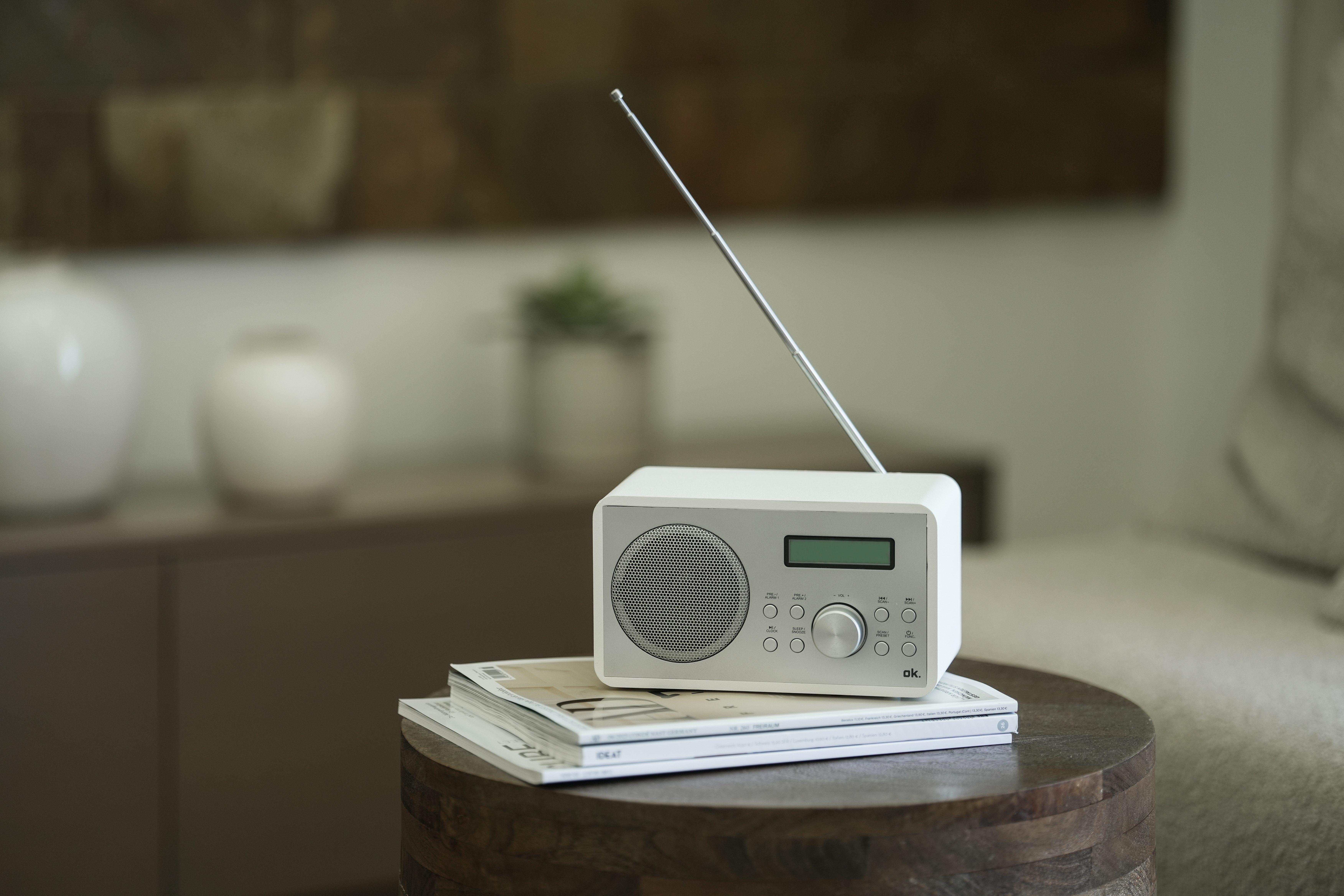 Ein weißes Radio mit Antenne steht auf Zeitschriften auf einem Holztisch. Der Hintergrund zeigt einen verschwommenen Raum.