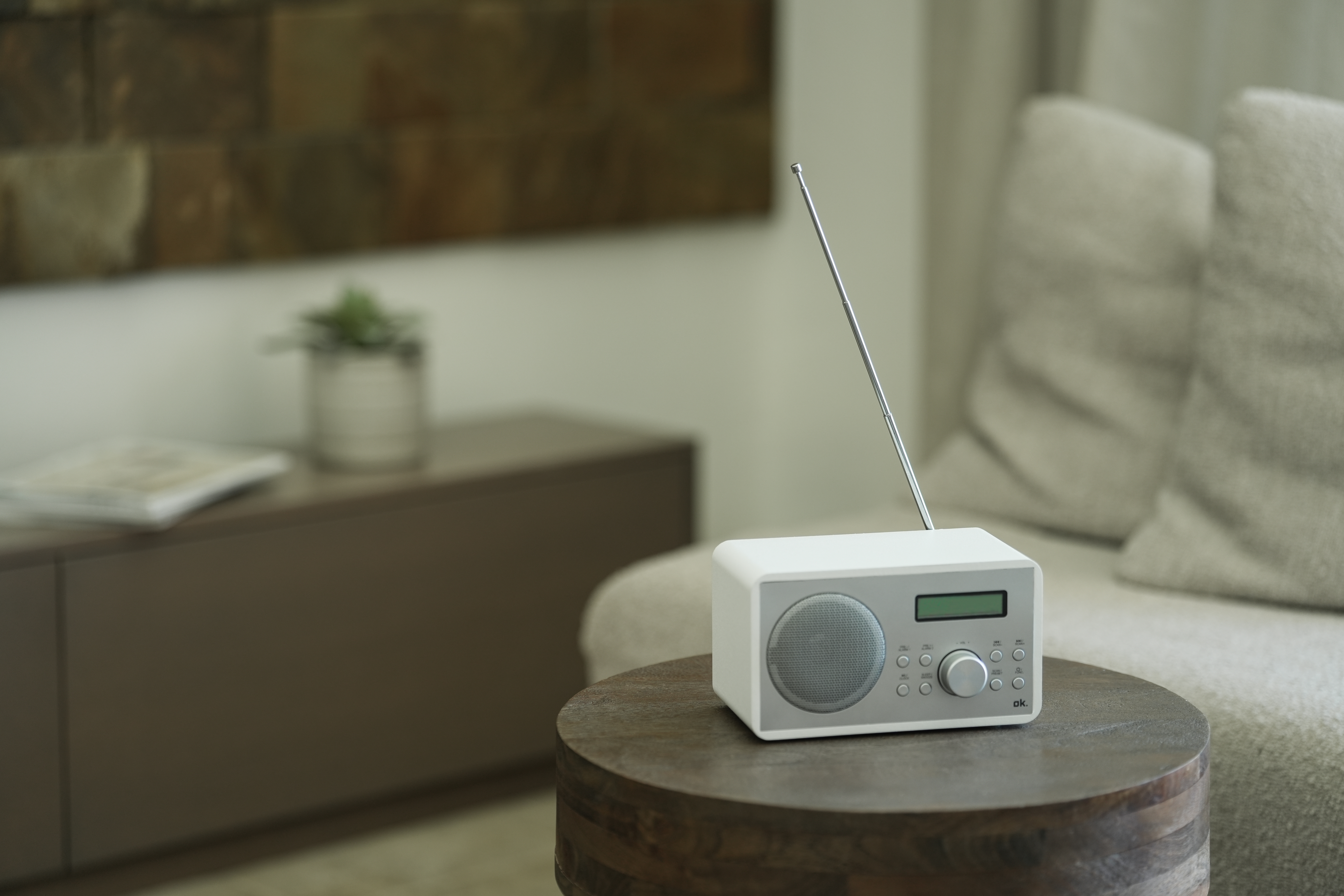 Ein weißes Radio mit Antenne steht auf einem Holztisch. Im Hintergrund sind ein Sofa und ein Schrank.