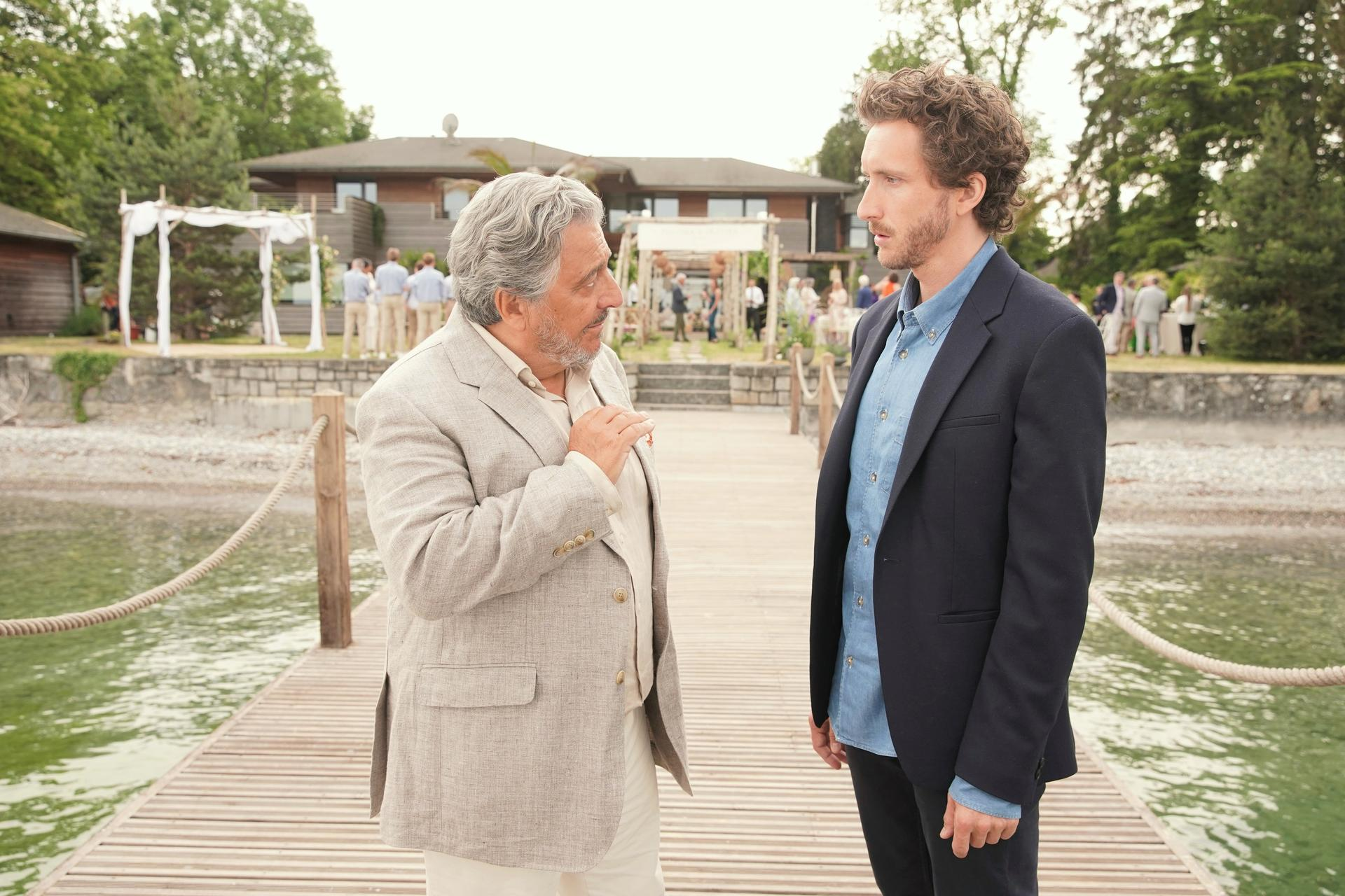 Zwei Männer auf einem Steg am Wasser, im Hintergrund ein Haus und Gäste.
