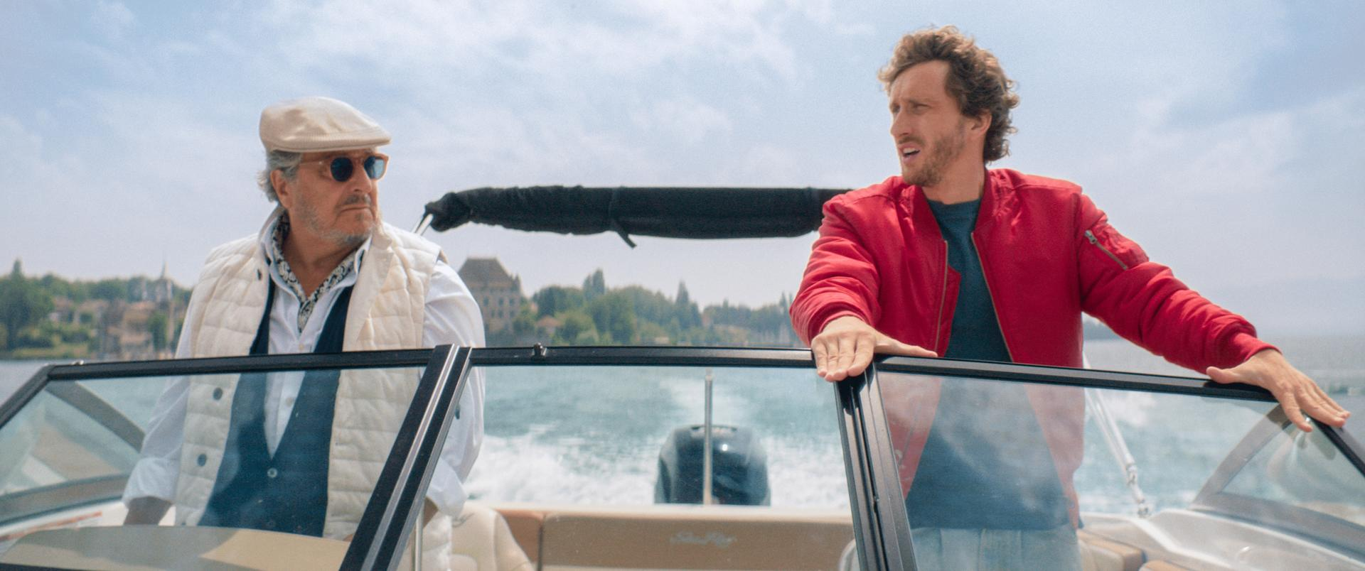 Zwei Männer auf einem Boot, einer trägt eine rote Jacke. Im Hintergrund Gebäude und Bäume.
