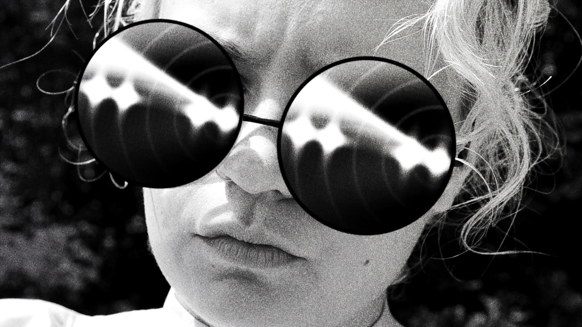 Schwarz-Weiß-Porträt einer Person mit runder Sonnenbrille. Reflexionen in den Gläsern. Haare im Hintergrund. Fokus auf das Gesicht.