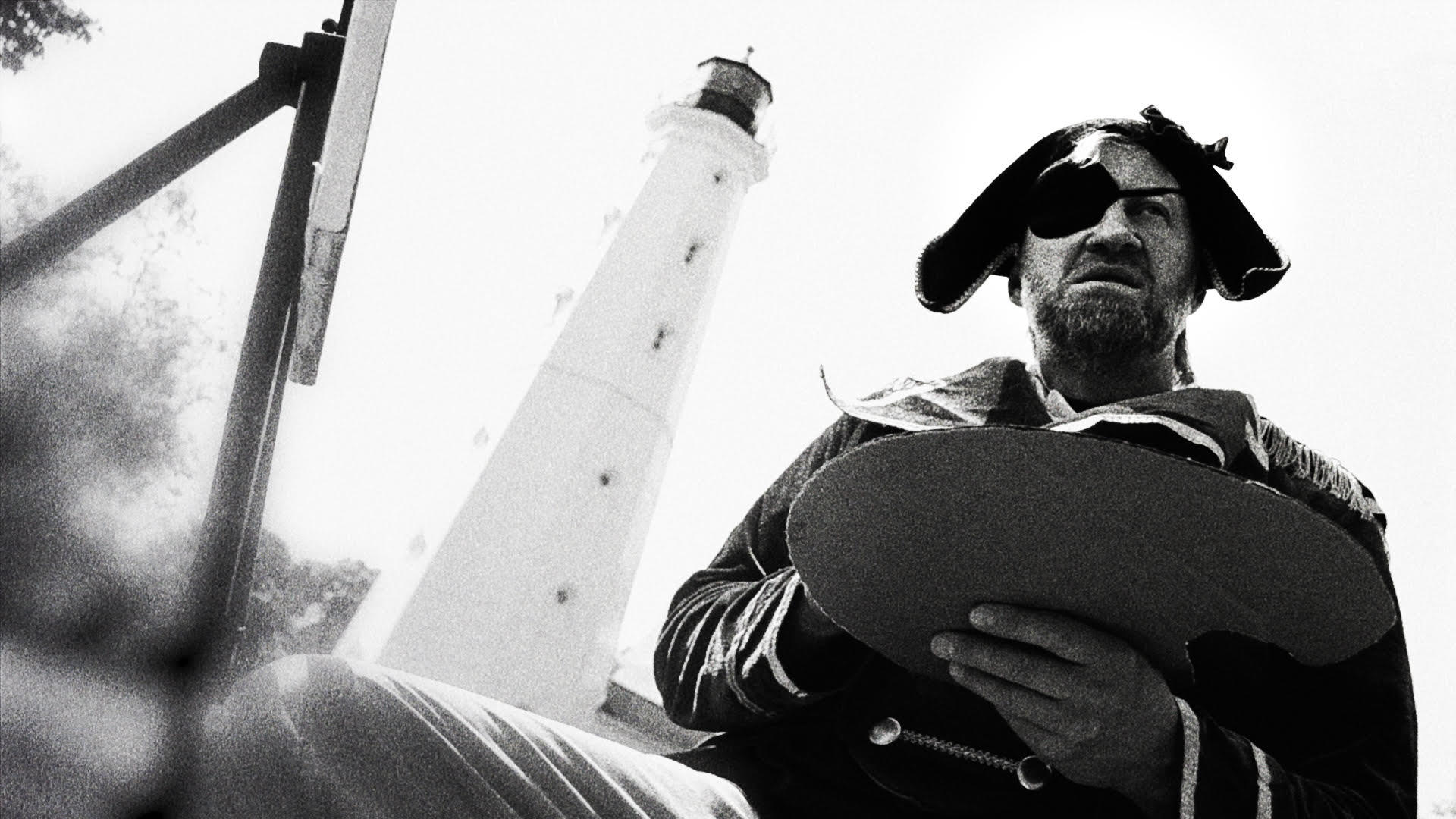 Ein Mann mit Augenklappe und Hut hält eine Palette vor einem Leuchtturm.
