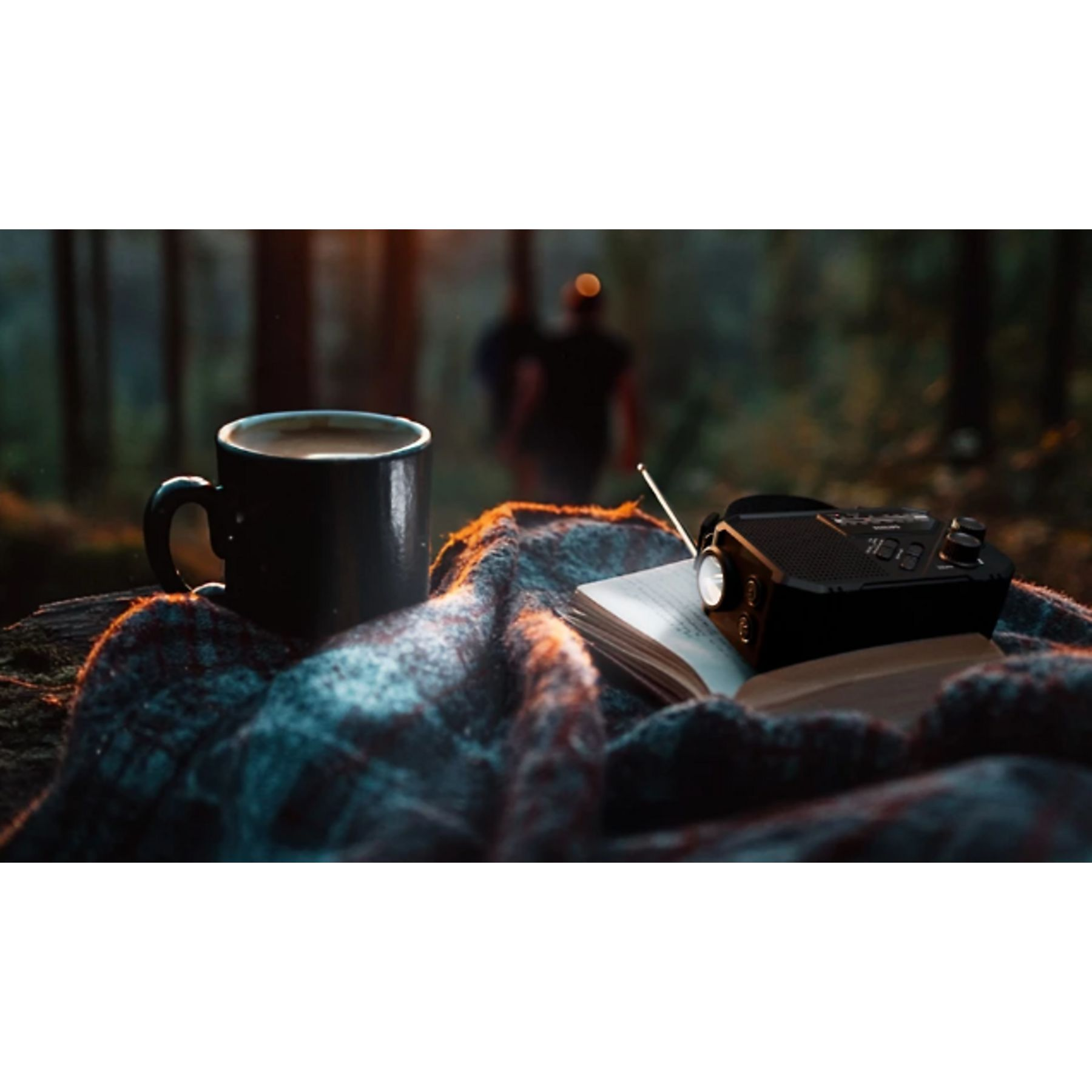 Une tasse de café, une radio et un livre sur une couverture dans une forêt. Deux personnes floues en arrière-plan.