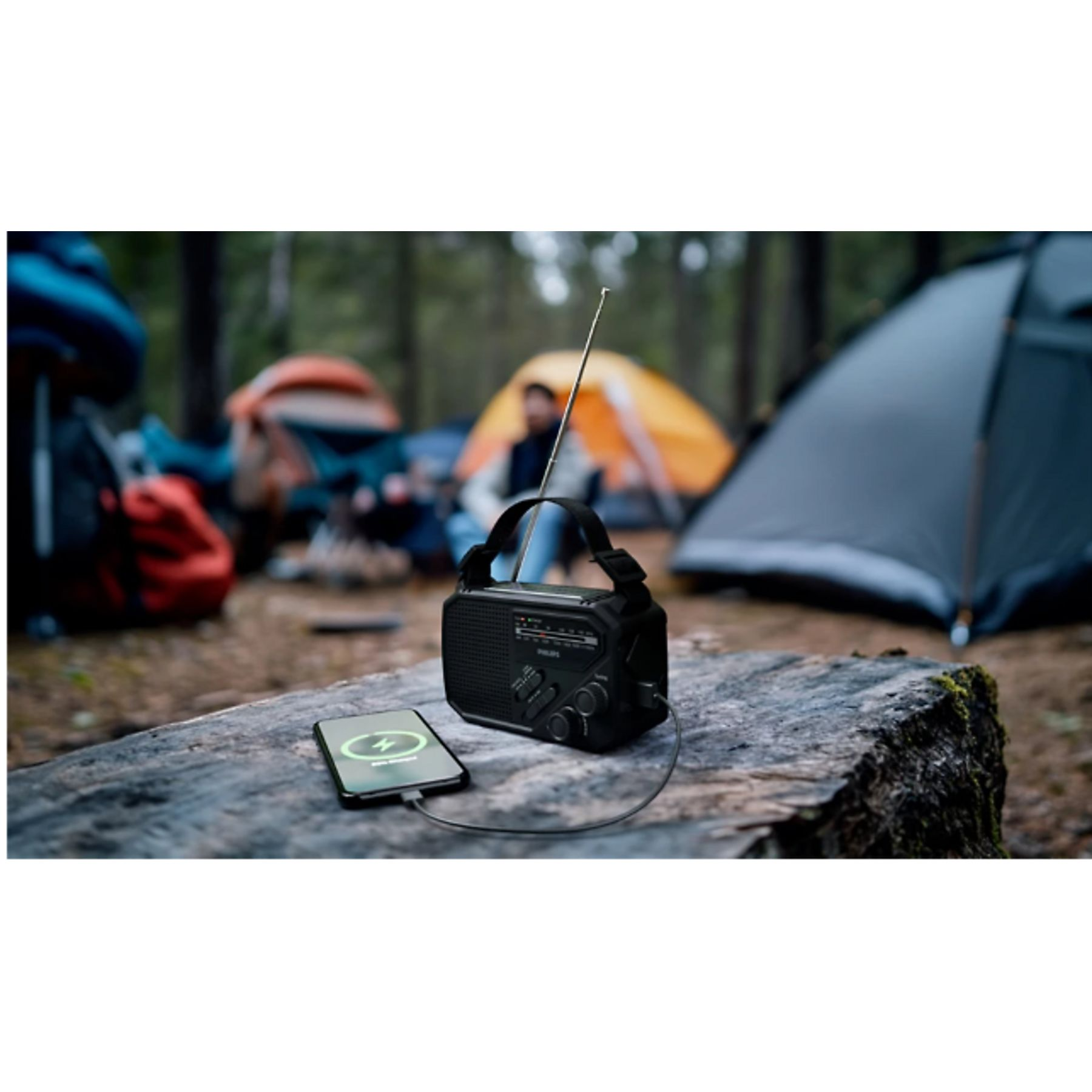 Une radio noire et un téléphone sont sur une bûche. Le téléphone se charge. Des tentes et des personnes sont en arrière-plan.