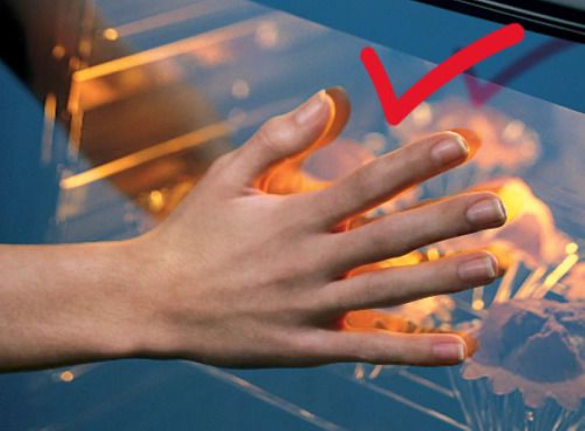 Ręka sprawdza piekarnik. Czerwony haczyk znajduje się na szkle. Wnętrze piekarnika jest widoczne z jedzeniem i światłem.