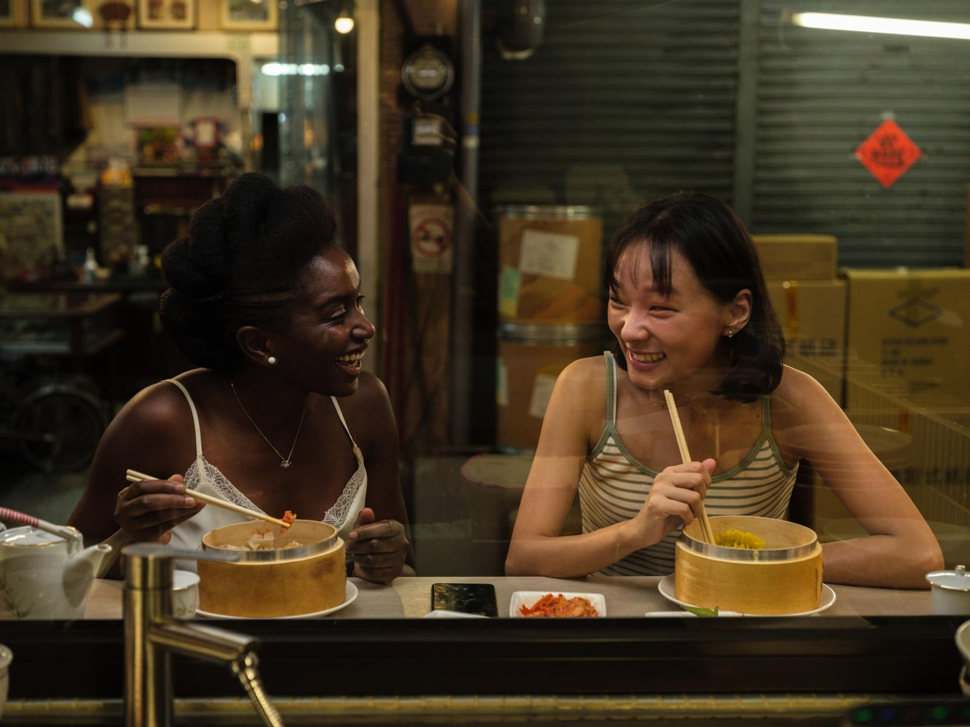 Zwei Frauen lachen beim Essen von Dim Sum. Sie sitzen an einem Tisch mit Essen in Bambusdämpfern. Der Hintergrund zeigt ein Restaurant.