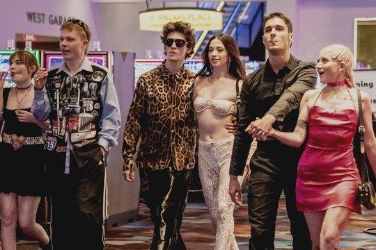 Sechs Personen gehen in einem Casino. Einer raucht eine Zigarette. Sie tragen verschiedene Outfits. Im Hintergrund sind Casino-Schilder und Spielautomaten zu sehen.
