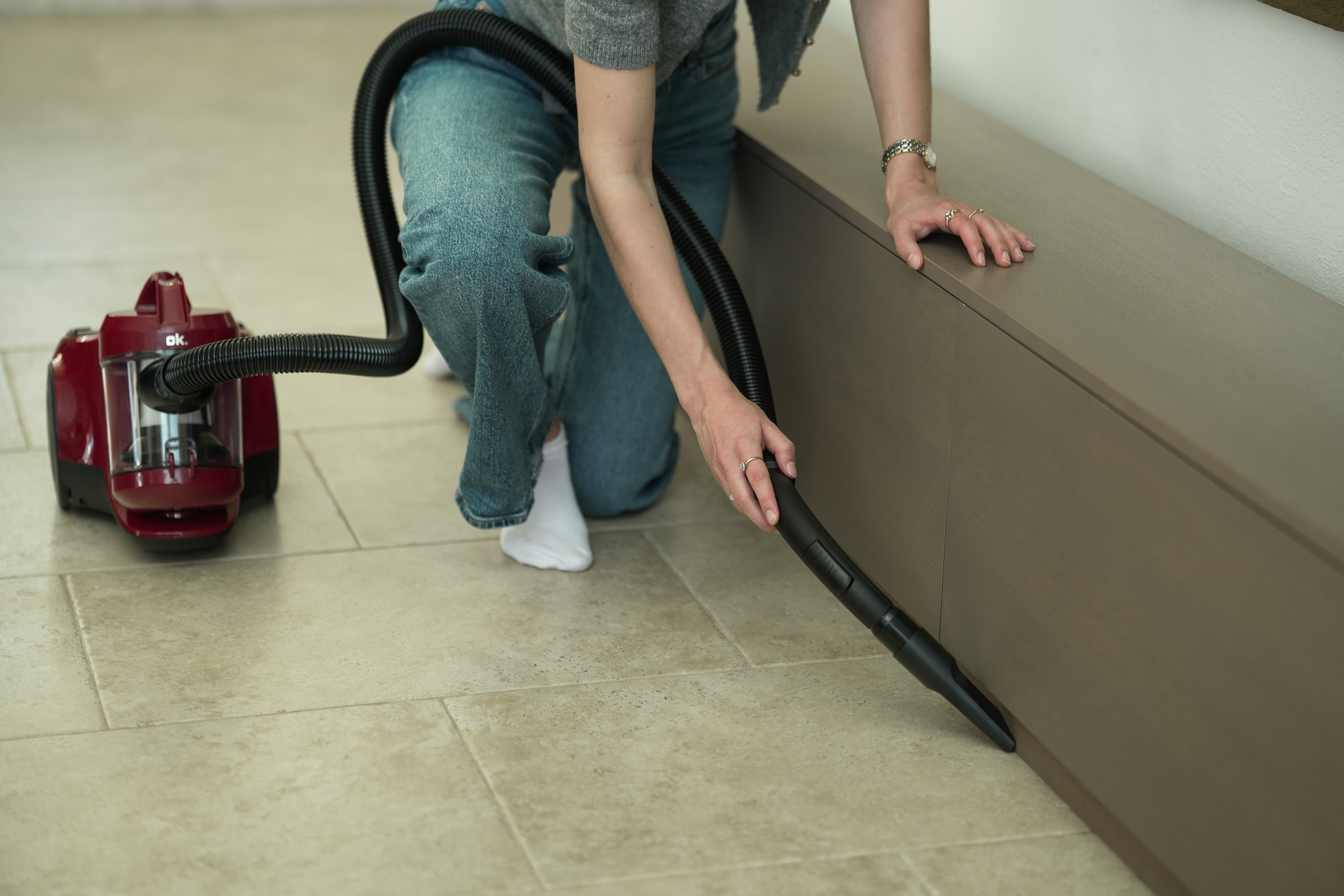 Eine Person saugt in der Nähe eines Schranks mit einem roten Staubsauger. Die Person trägt Jeans und einen grauen Pullover.