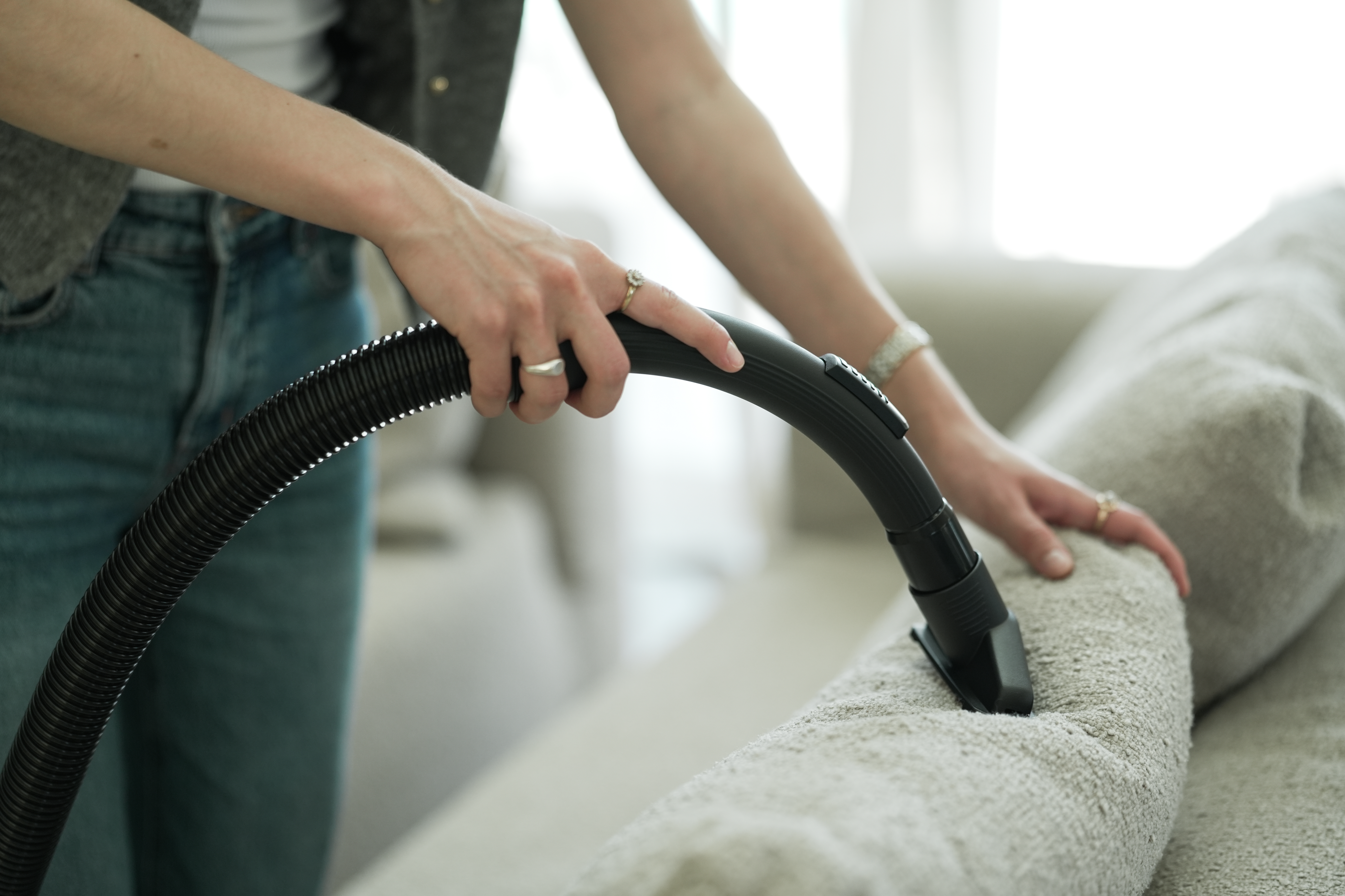 Eine Person saugt ein hellgraues Sofa mit einem schwarzen Staubsauger. Die Person trägt Jeans und eine graue Weste.