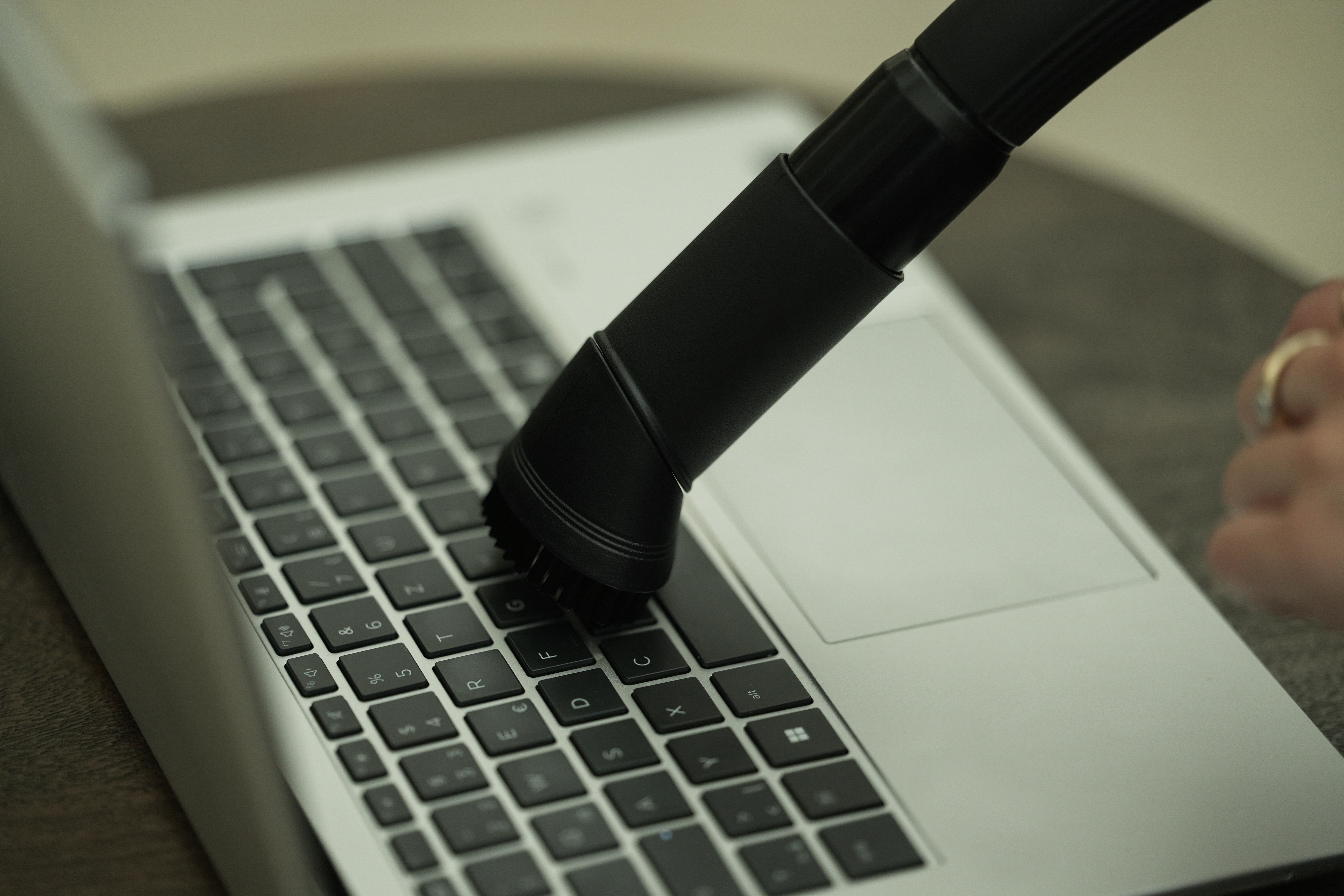 Eine Laptop-Tastatur wird mit einem schwarzen Staubsauger gereinigt. Eine Hand mit Ring ist sichtbar.