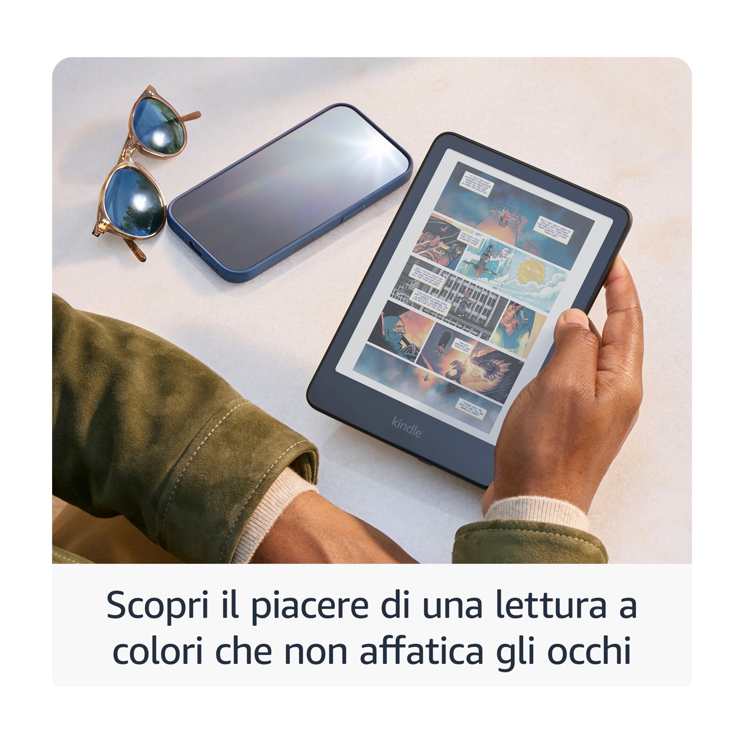Una persona tiene in mano un Kindle che mostra un fumetto. Occhiali da sole e telefono accanto. La persona indossa una giacca verde.