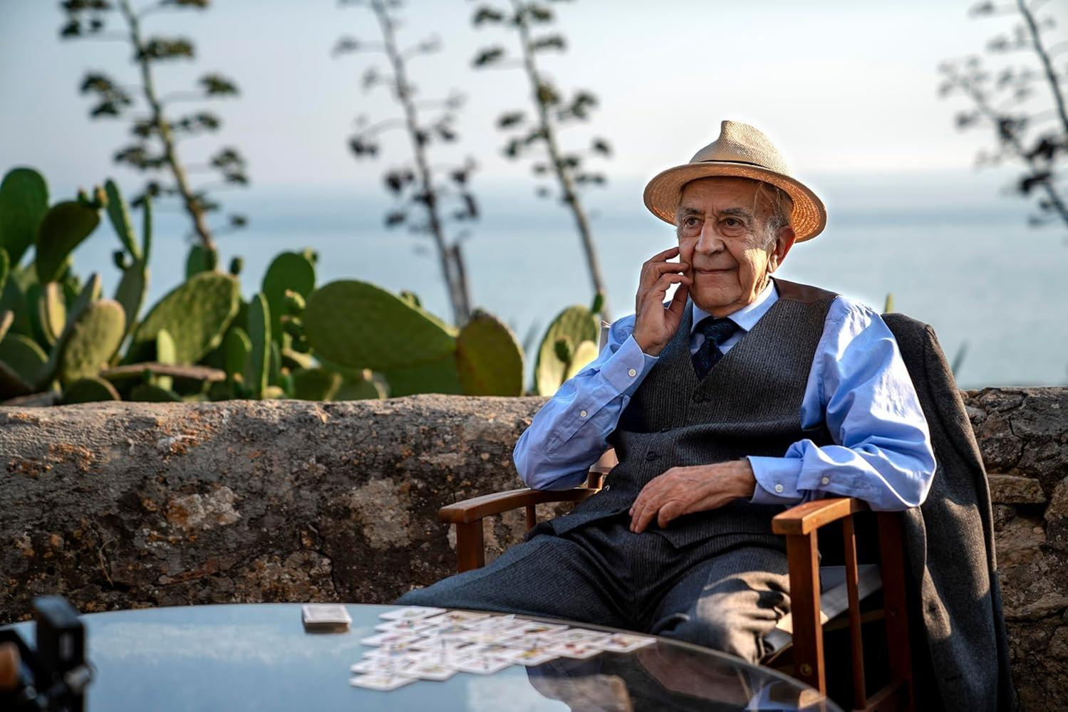 Ein älterer Mann mit Hut sitzt und berührt sein Gesicht. Karten auf einem Tisch, Meer und Pflanzen im Hintergrund. Er trägt eine Weste und ein blaues Hemd.