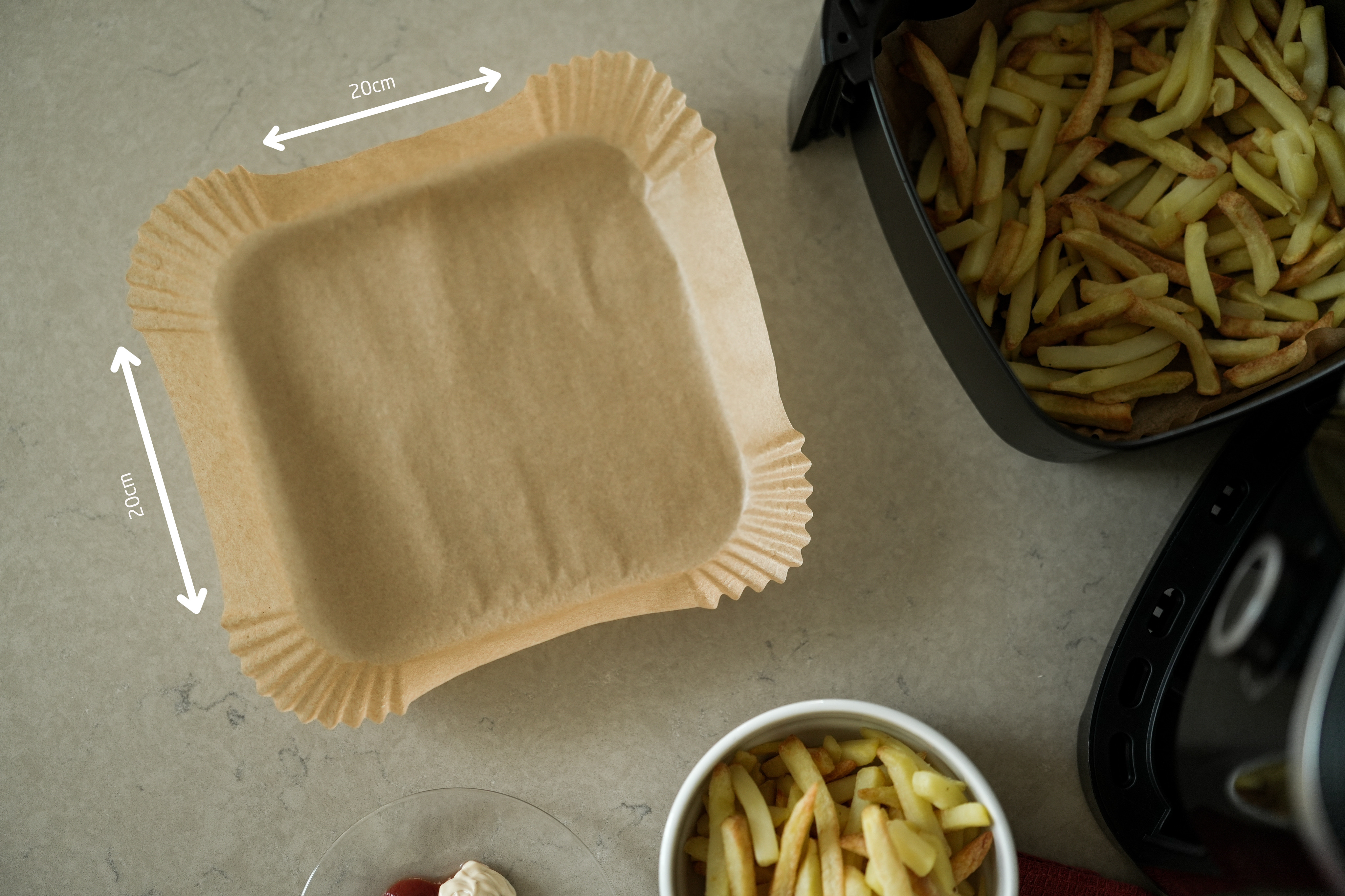Ein Backpapier, eine Heißluftfritteuse mit Pommes, eine Schüssel Pommes und Gewürze auf einer Arbeitsplatte.
