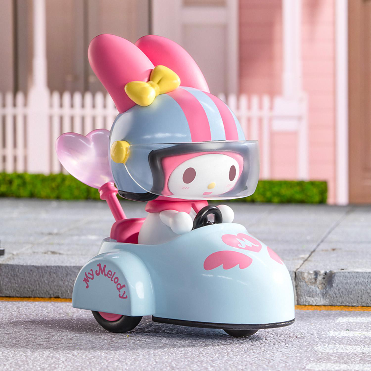 My Melody Spielfigur in einem blauen Auto mit rosa Details. Sie trägt einen Helm und eine Schleife und fährt auf einer Straße mit Haus im Hintergrund.