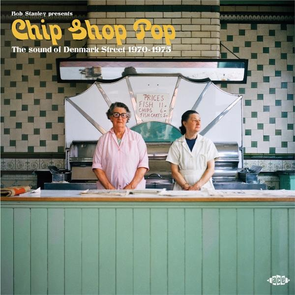 Zwei Frauen stehen hinter der Theke eines Imbissladens. Ein Schild zeigt Preise. Der Laden hat grüne Täfelung und geflieste Wände. Das Schild sagt "Chip Shop Pop".