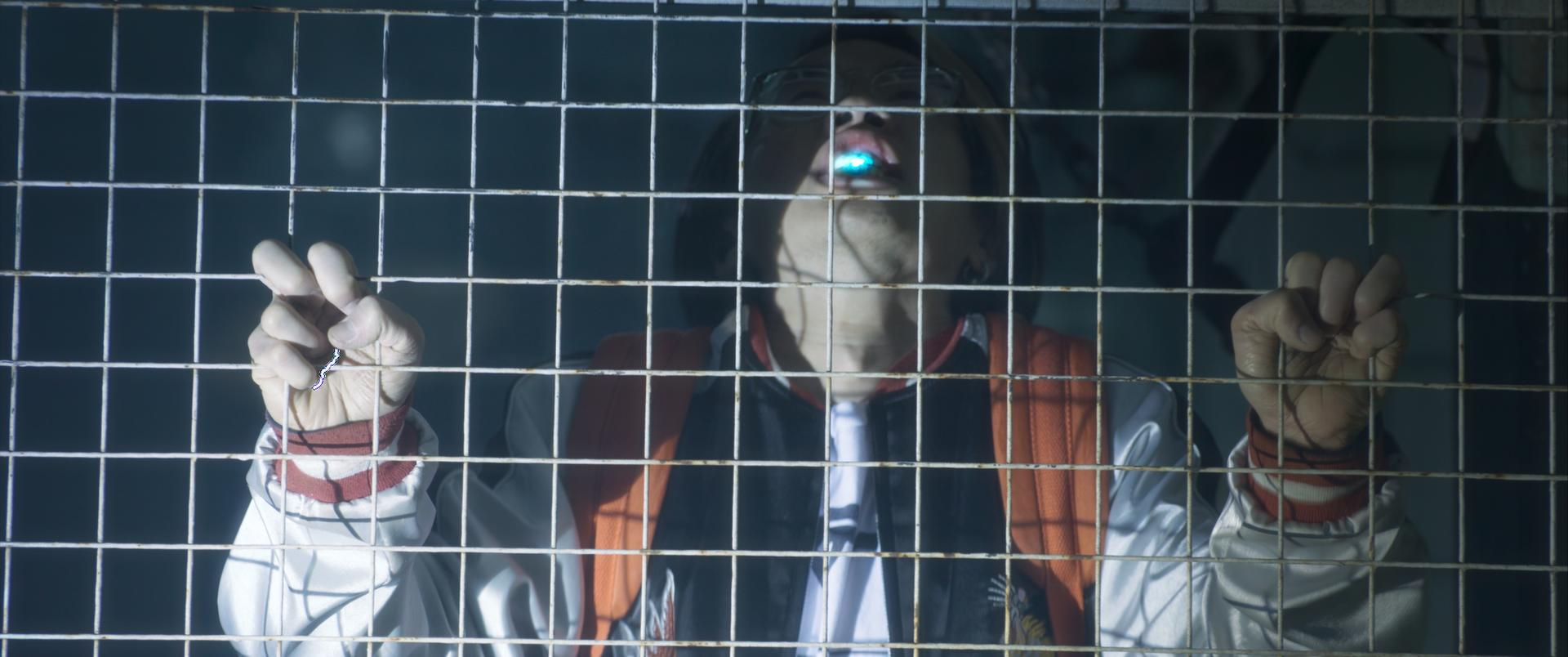 Person hinter einem Metallzaun. Sie tragen eine Brille und eine Jacke. Ein blaues Leuchten ist in ihrem Mund. Hände greifen den Zaun.