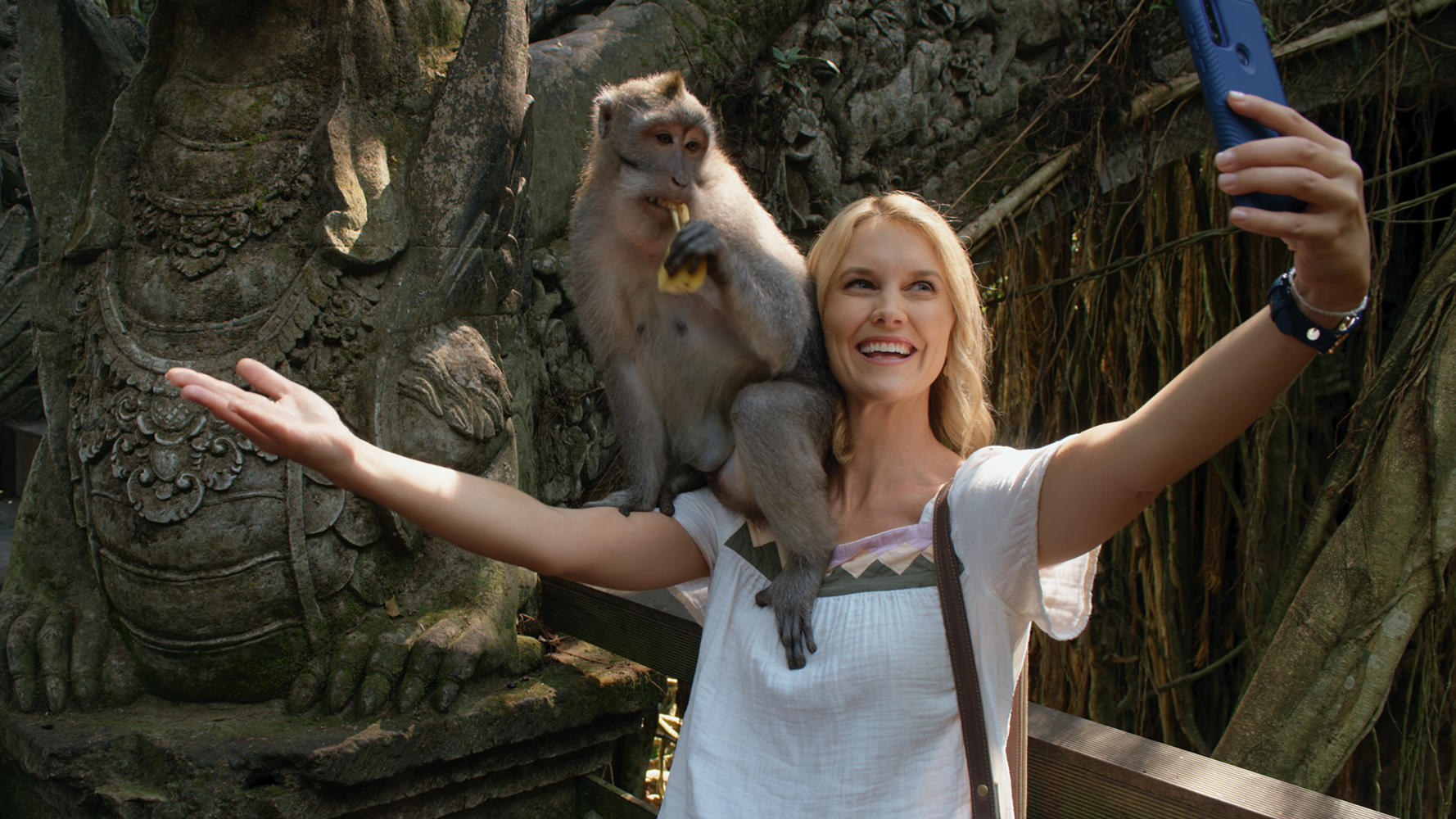 Eine Frau macht ein Selfie mit einem Affen auf ihrer Schulter. Der Affe isst eine Banane. Steinschnitzerei im Hintergrund.