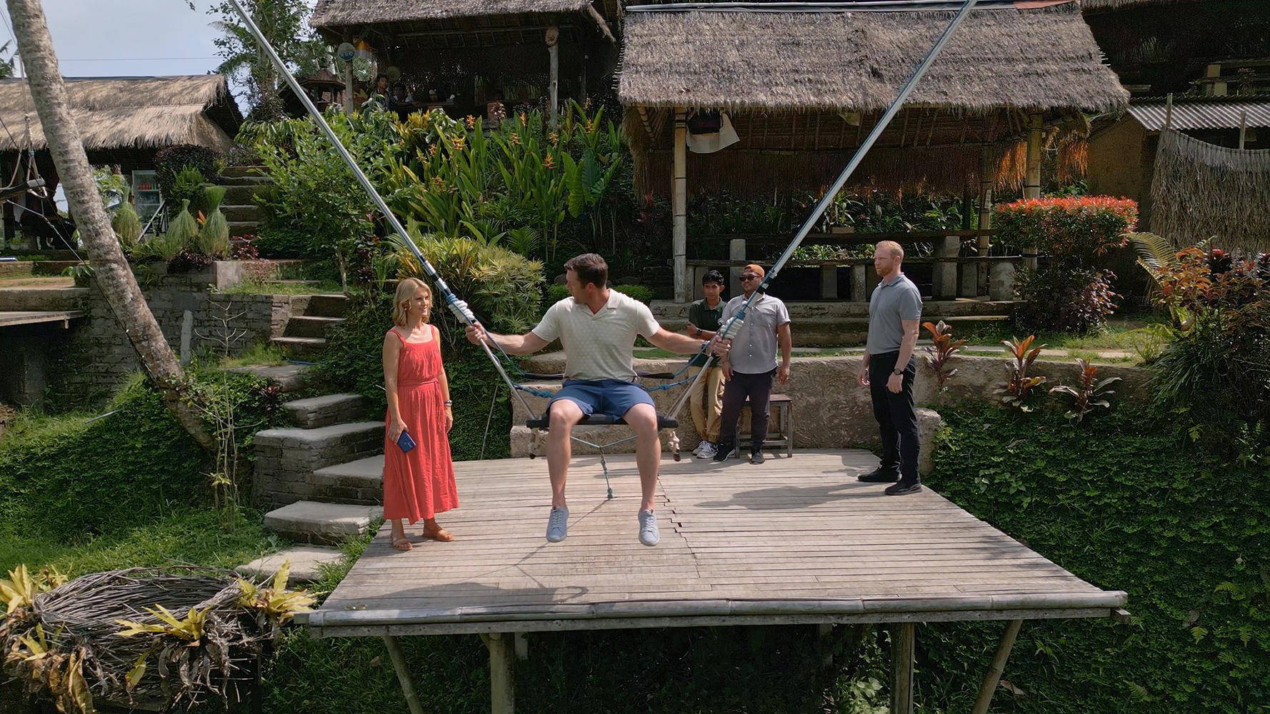Mann auf einer Schaukel, zwei Frauen, drei Männer stehen auf einem Holzdeck. Üppige grüne Vegetation und Gebäude mit Strohdächern im Hintergrund.