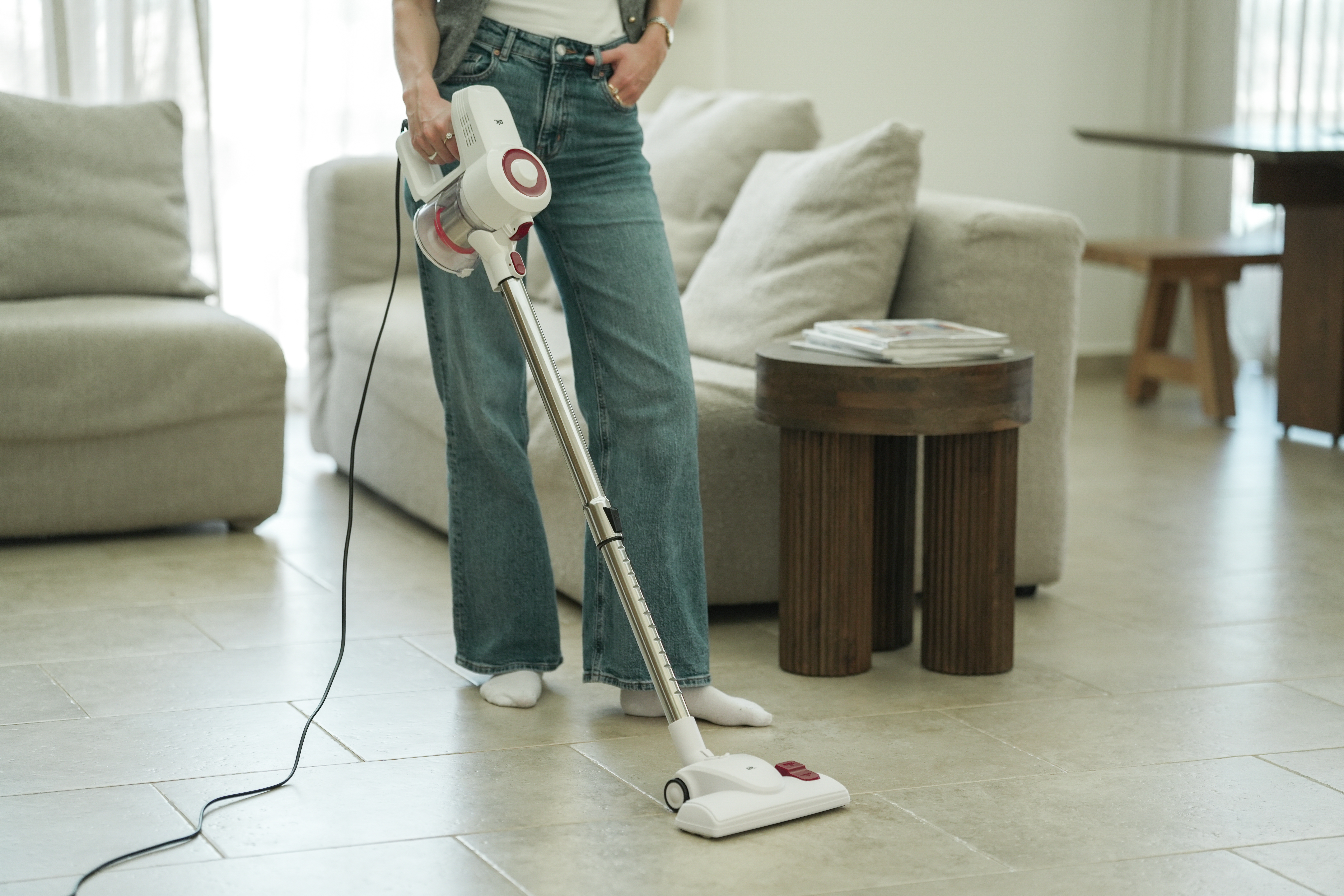 Eine Person in Jeans saugt mit einem weiß-roten Staubsauger einen gefliesten Boden. Im Hintergrund sind ein Sofa und ein Tisch.