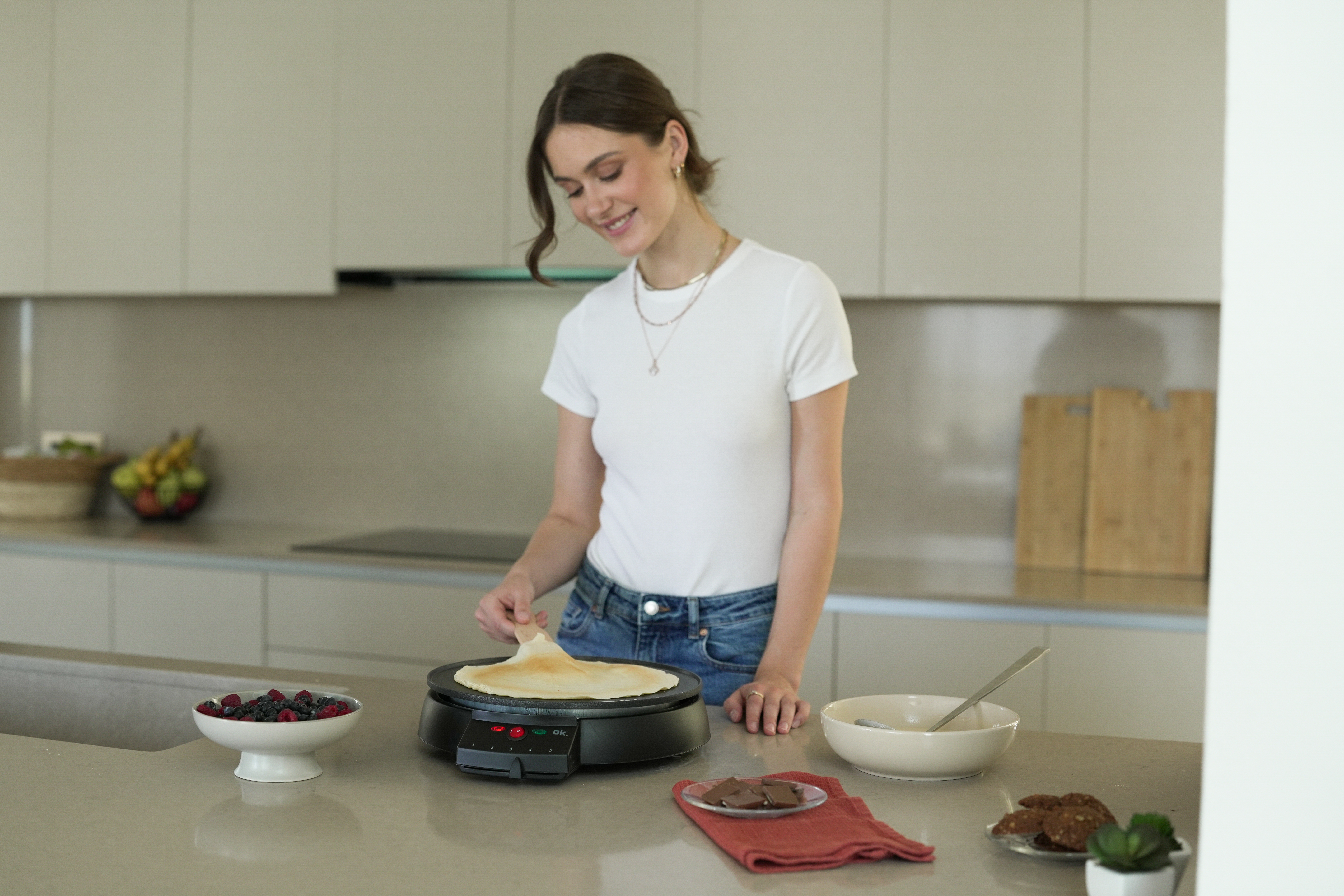 Frau backt einen Pfannkuchen auf einer Grillplatte in einer modernen Küche. Beeren, Kekse und eine Schüssel mit Teig sind sichtbar.