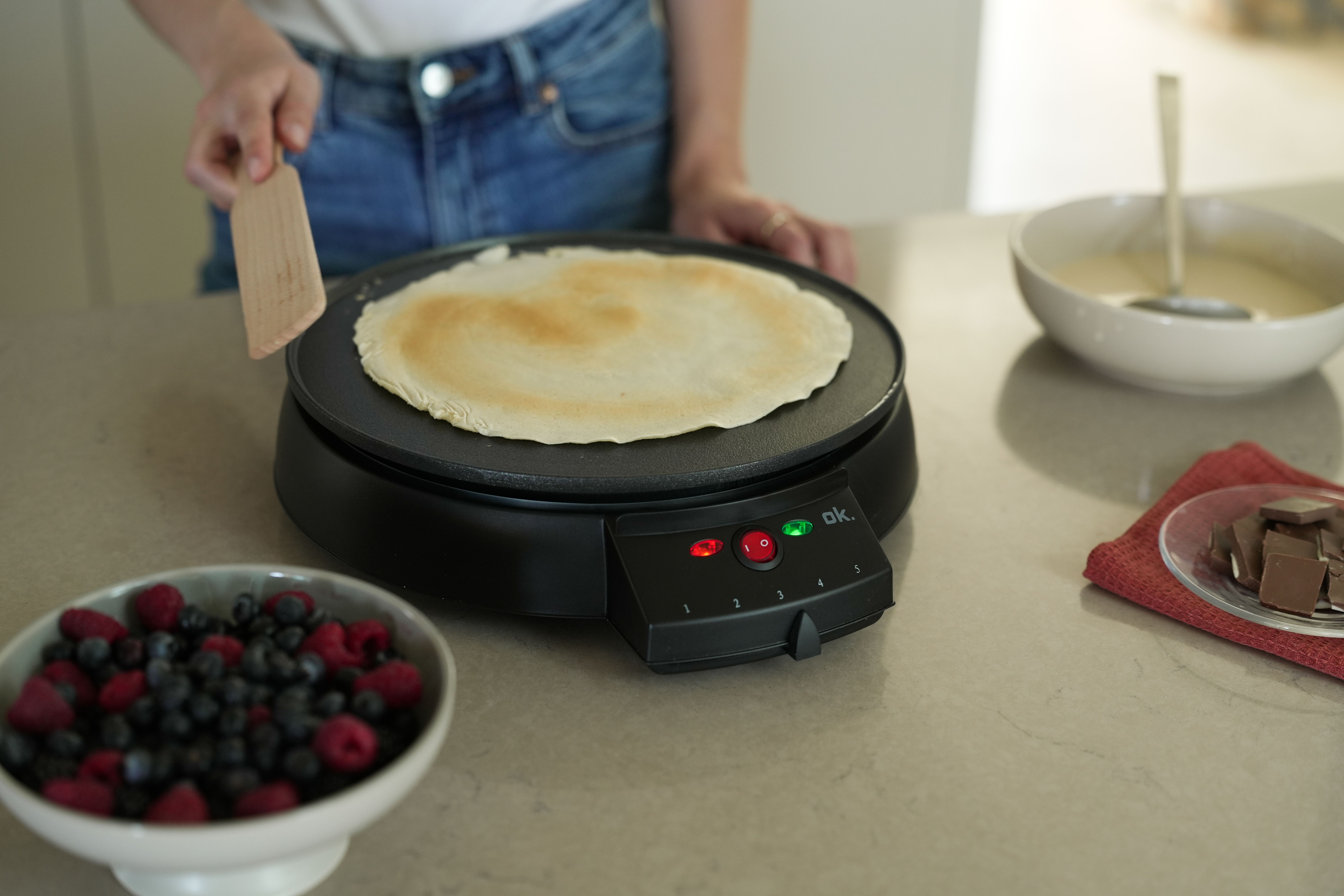 Eine Person macht einen Crêpe auf einem Elektrogrill. Beeren, Schokolade und Teig sind in der Nähe. Ein Holzspatel wird verwendet.
