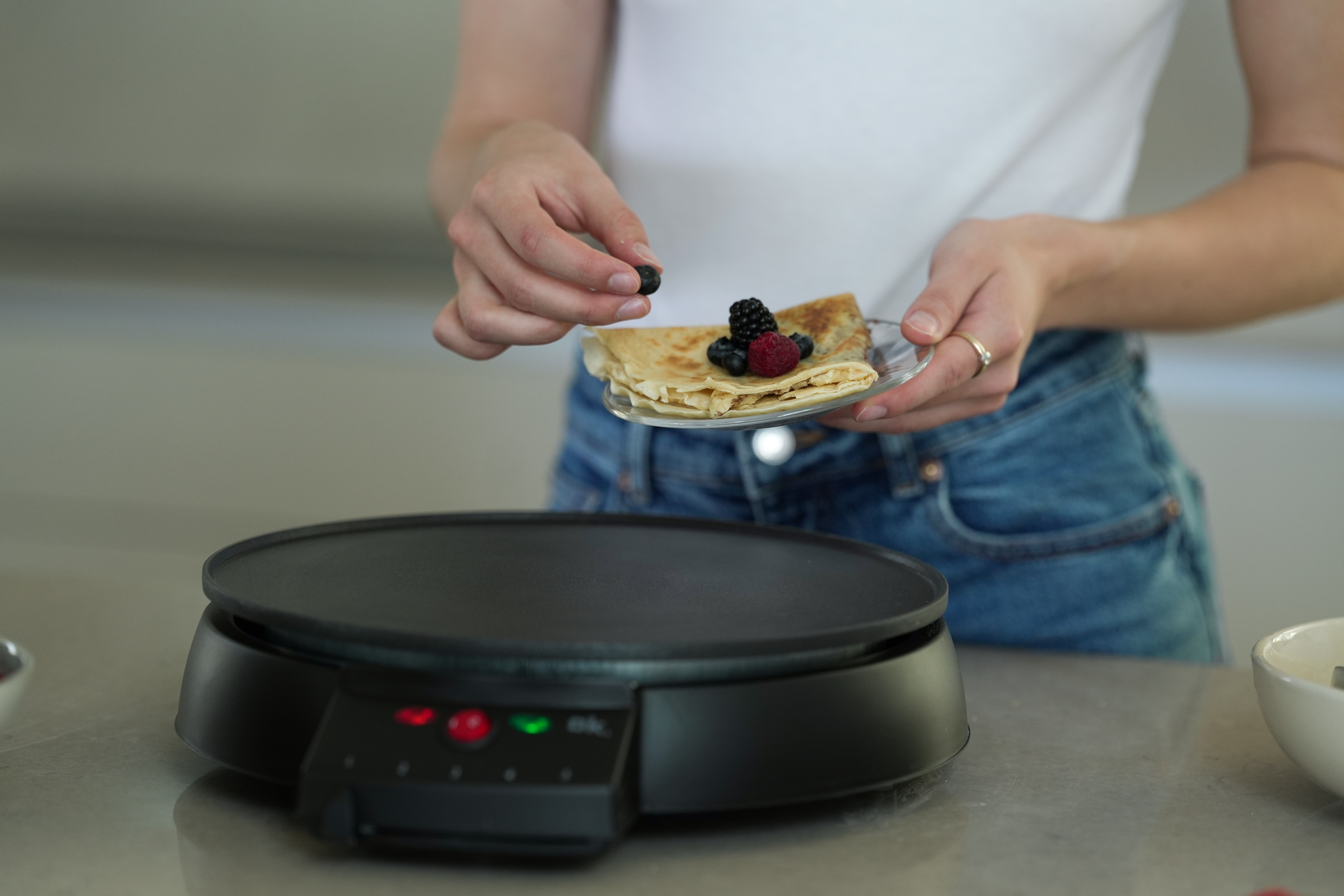 Eine Person fügt Blaubeeren zu einem Crêpe mit Beeren auf einem Teller hinzu. Auf dem Tisch steht ein schwarzer Crêpe-Maker.
