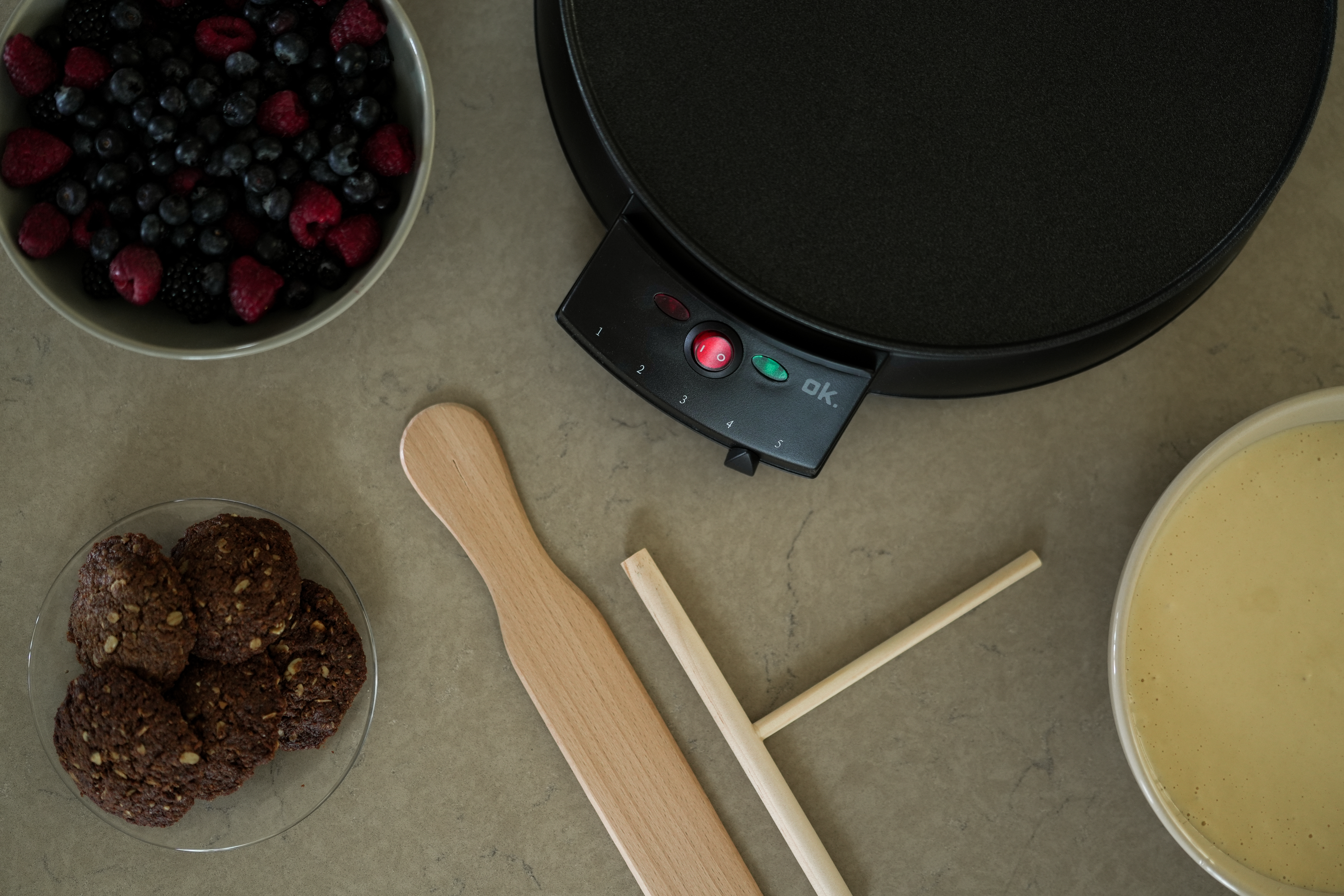 Ein Pfannkuchenmacher mit Zutaten wie Beeren, Keksen und Teig. Holzutensilien sind vorhanden.