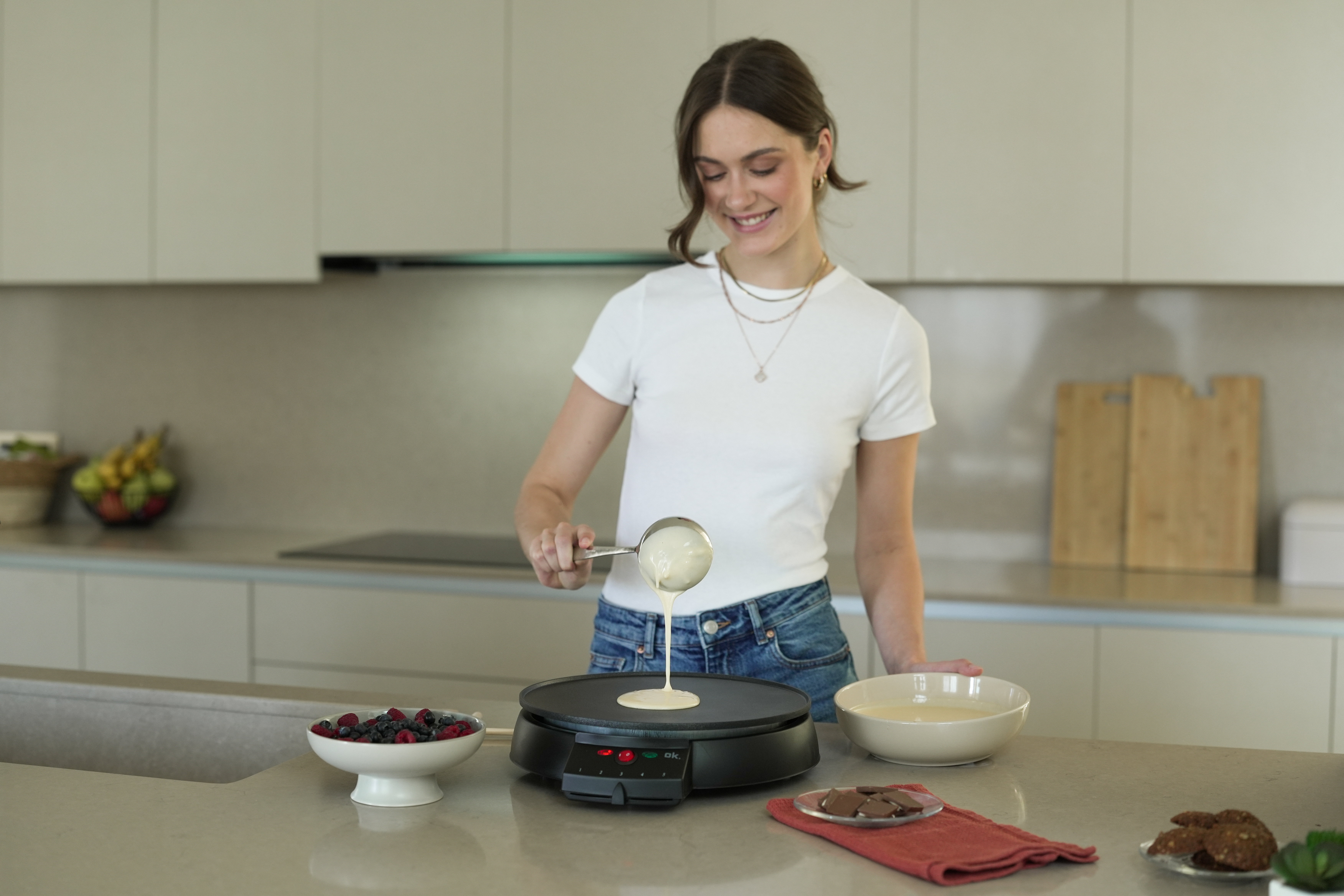Eine Frau gießt Teig auf einen Crêpes-Maker. Beeren, Kekse und Teig liegen auf der Theke.