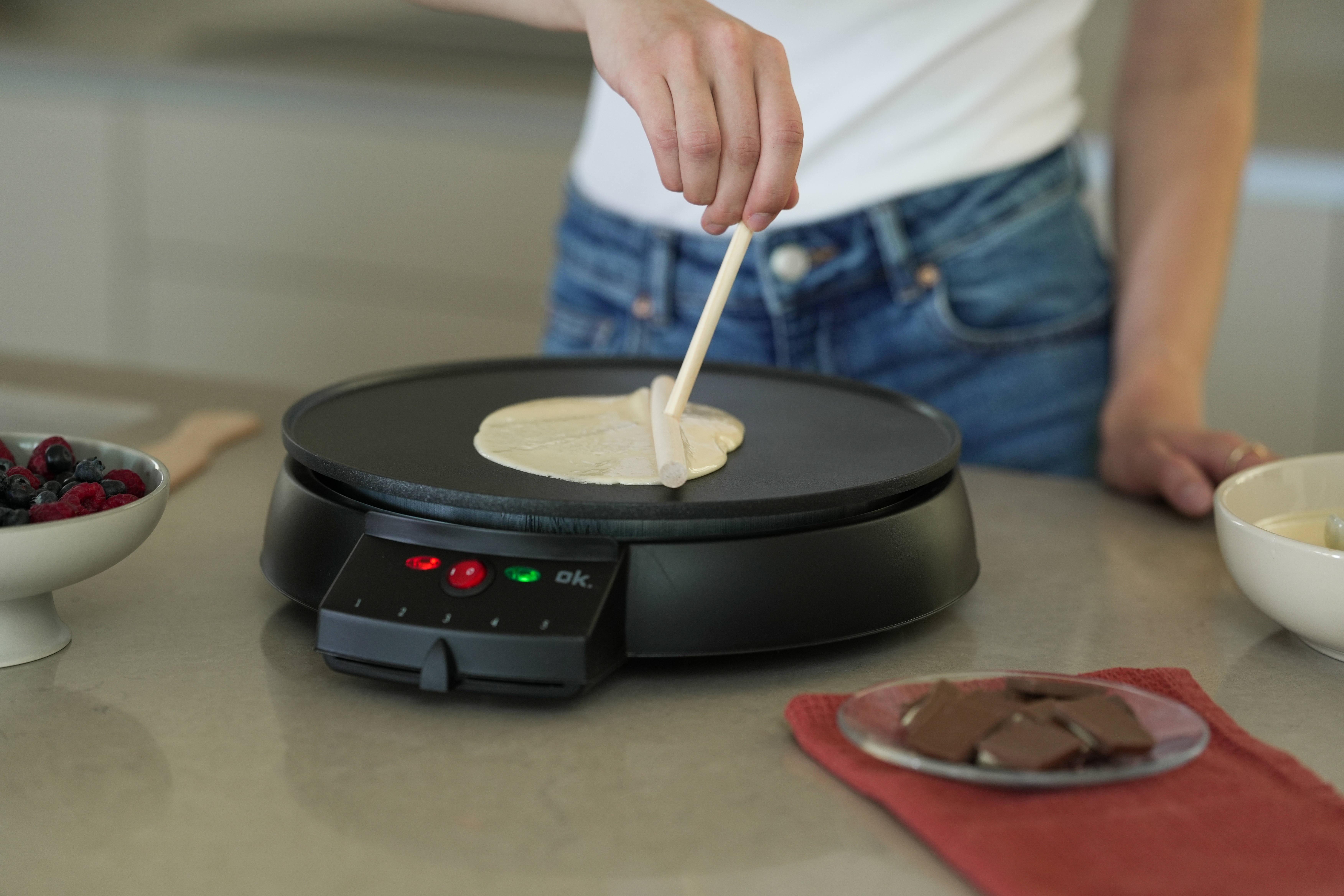 Eine Person macht einen Crêpe auf einem schwarzen Crêpe-Maker. Beeren und Schokolade liegen auf der Theke.