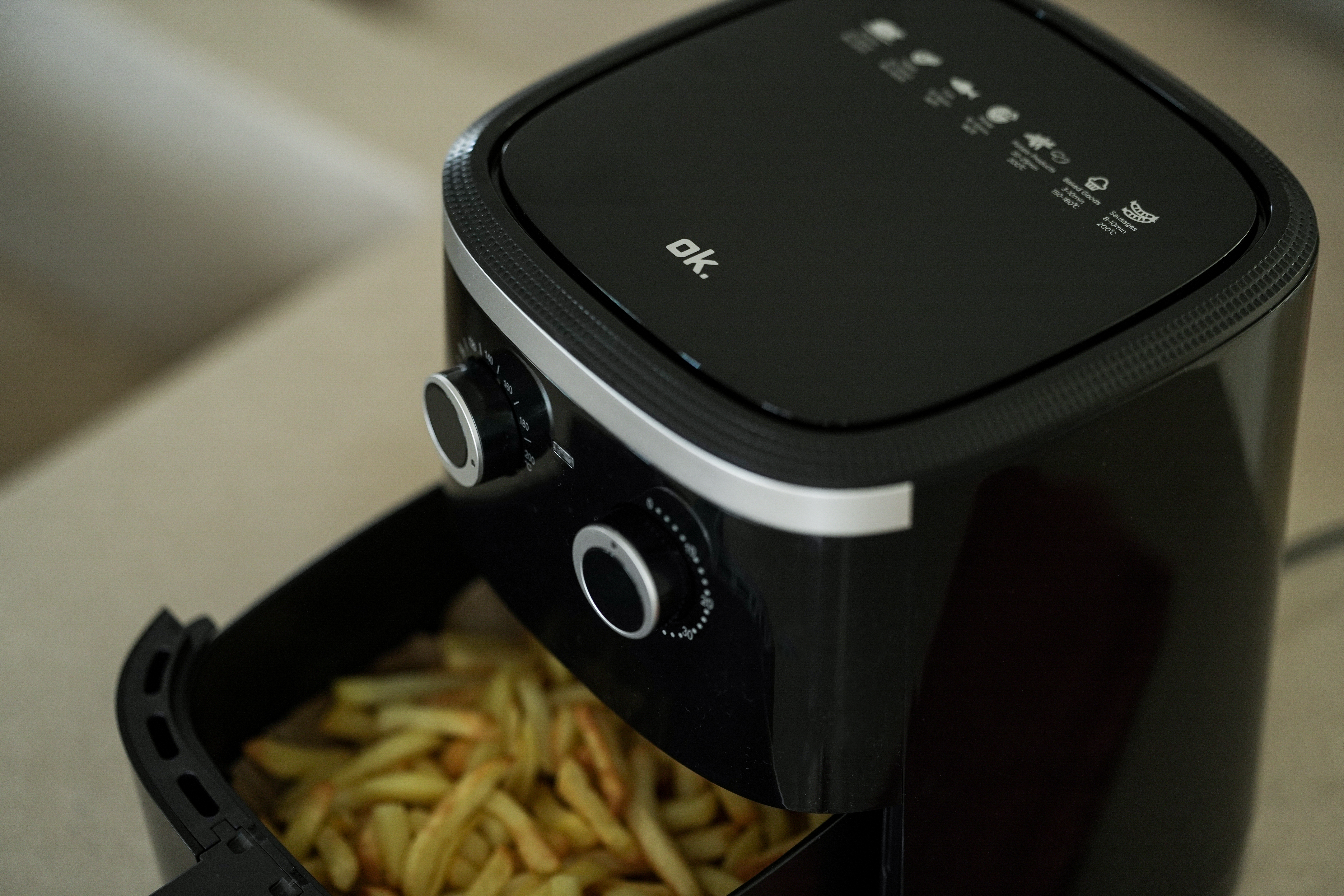 Schwarze Heißluftfritteuse mit Pommes Frites. Knöpfe und Einstellungen sind sichtbar. Der Markenname 'ok.' steht oben.