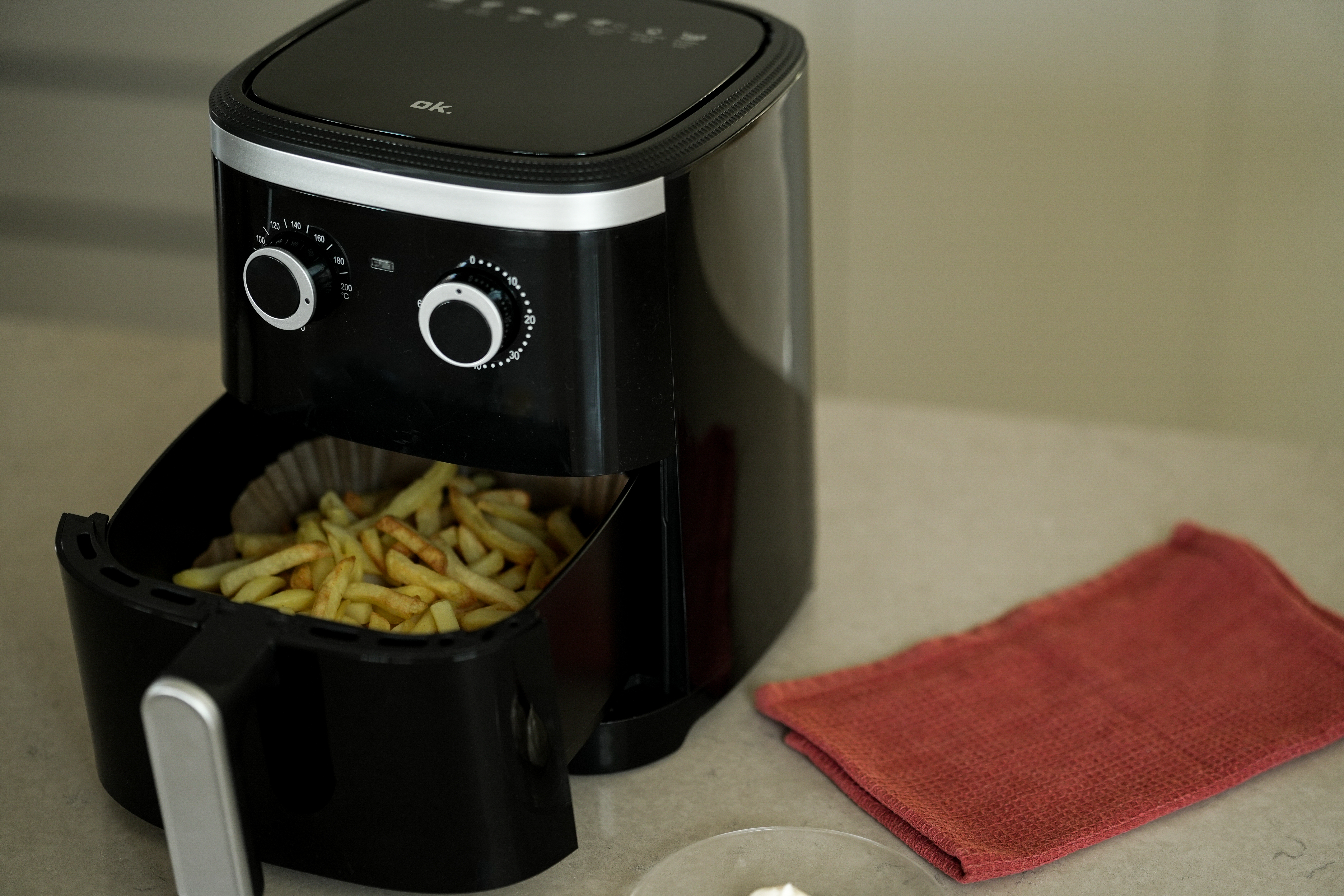 Ein schwarzer Airfryer mit Pommes Frites. Ein rotes Tuch und ein Gericht mit Sahne stehen auf dem Tisch.