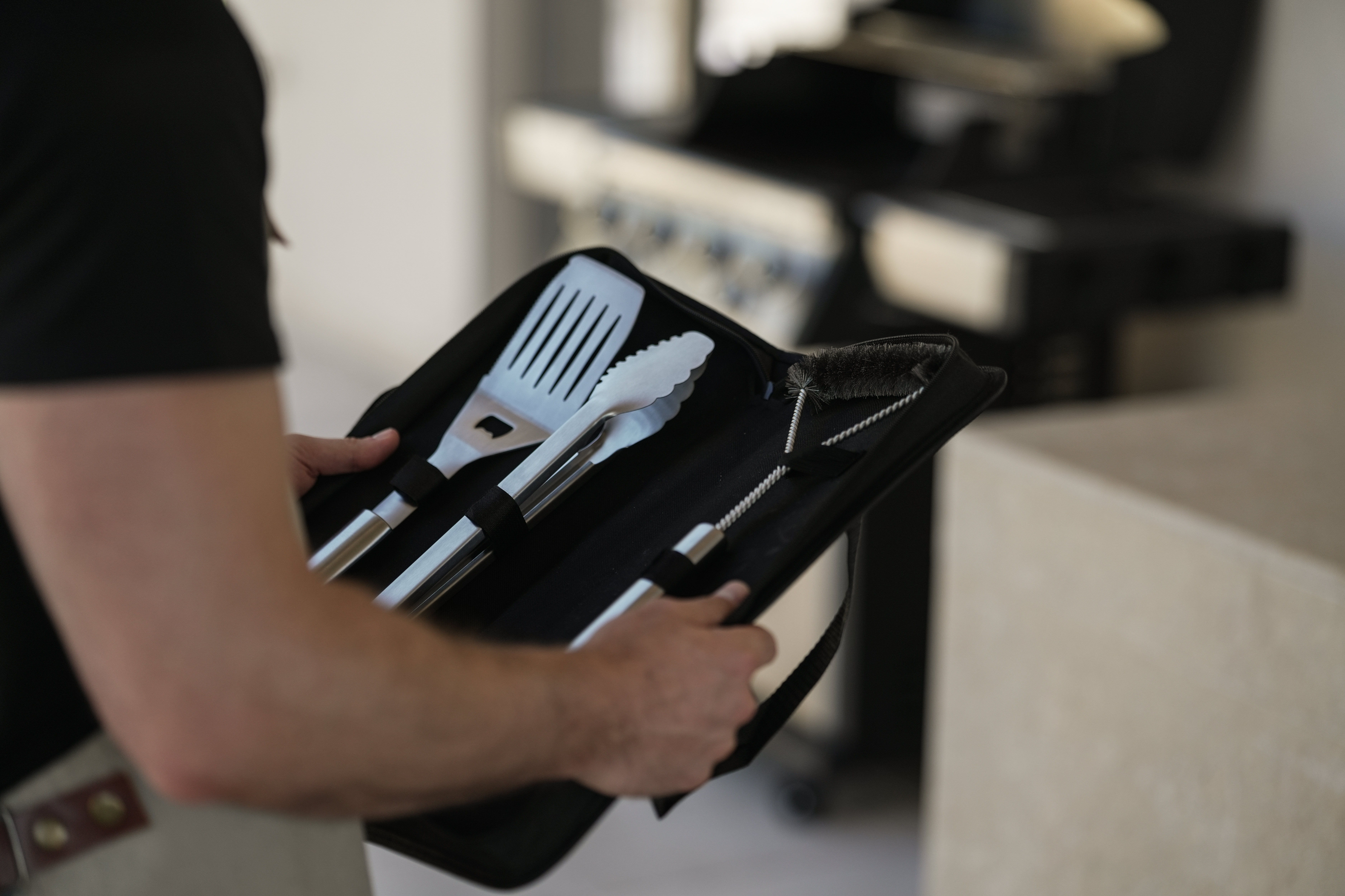 Eine Person hält einen schwarzen Koffer mit Grillwerkzeugen, darunter ein Spatel, eine Zange und eine Bürste, mit einem Grill im Hintergrund.