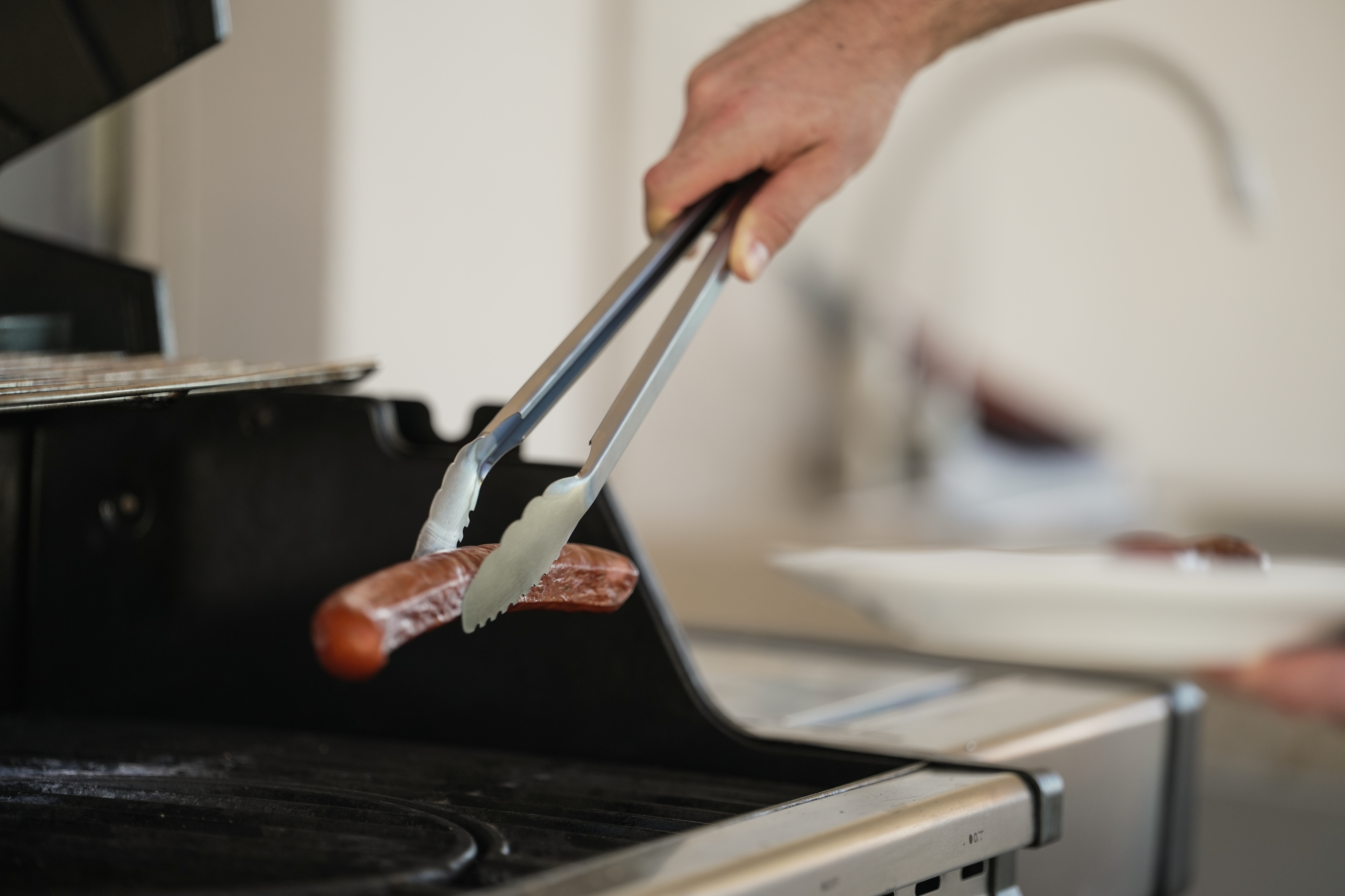 Eine Hand benutzt eine Zange, um eine Wurst auf einen Grill zu legen. Ein Teller ist im Hintergrund.