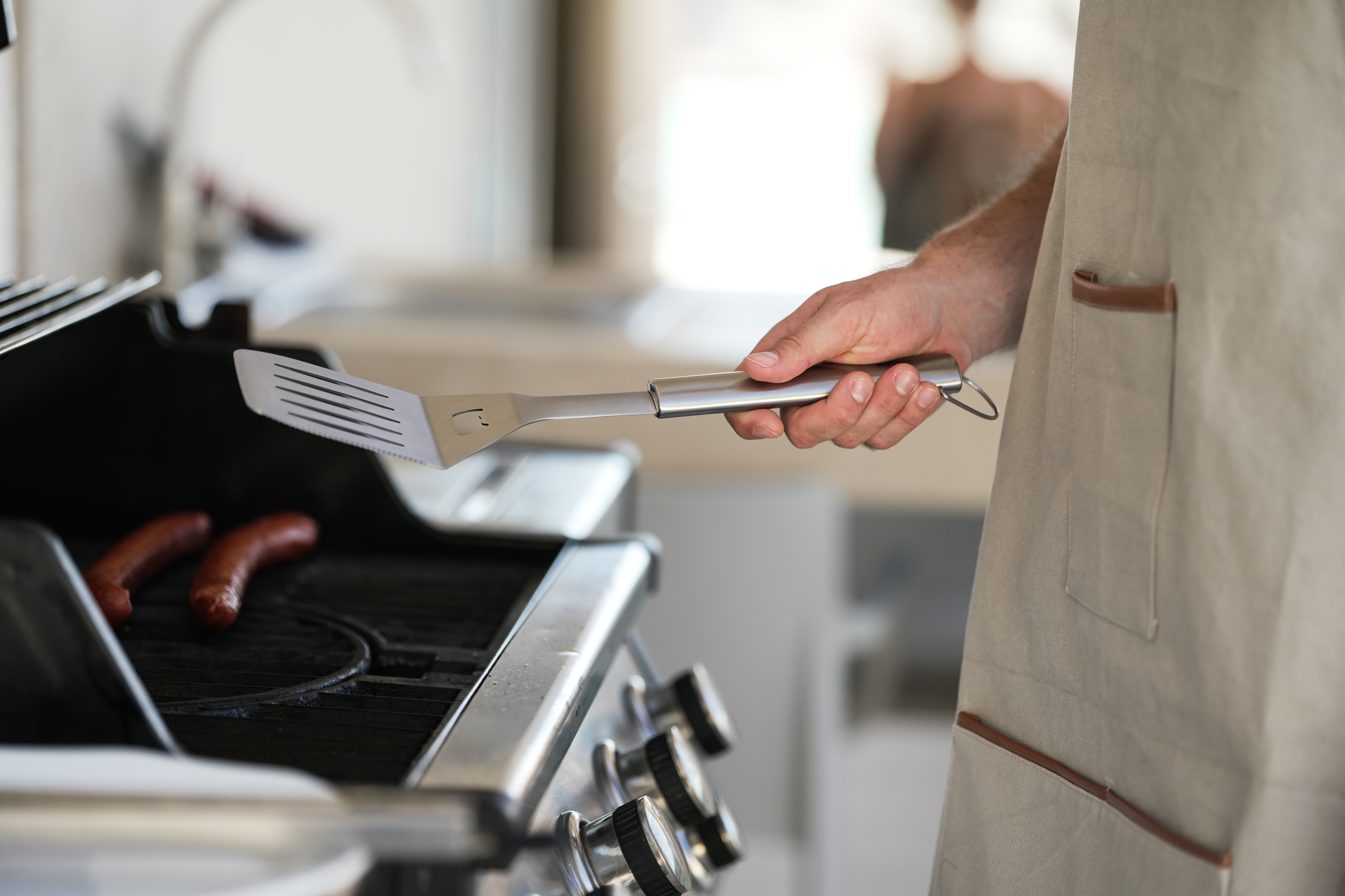 Eine Hand hält einen Spatel über einem Grill mit Würstchen. Eine Person mit Schürze steht daneben.