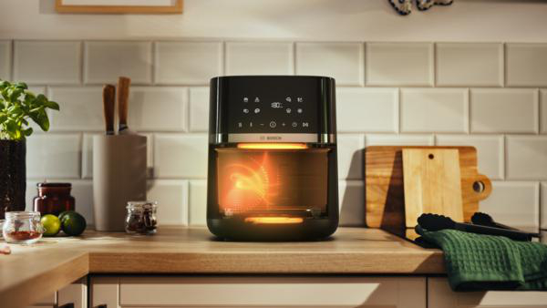 Ein Airfryer auf einer Holztheke. Dahinter befinden sich weiße Fliesen. Es gibt Limetten, Gläser, ein Schneidebrett und ein grünes Handtuch.