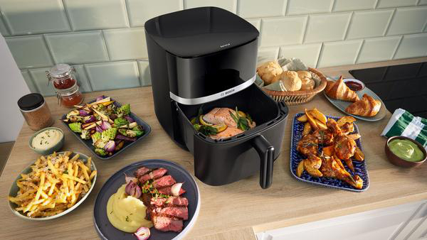 Ein schwarzer Airfryer mit verschiedenen Speisen: Pommes, Gemüse, Steak, Hähnchen, Brot und Croissants auf einer Holzoberfläche.