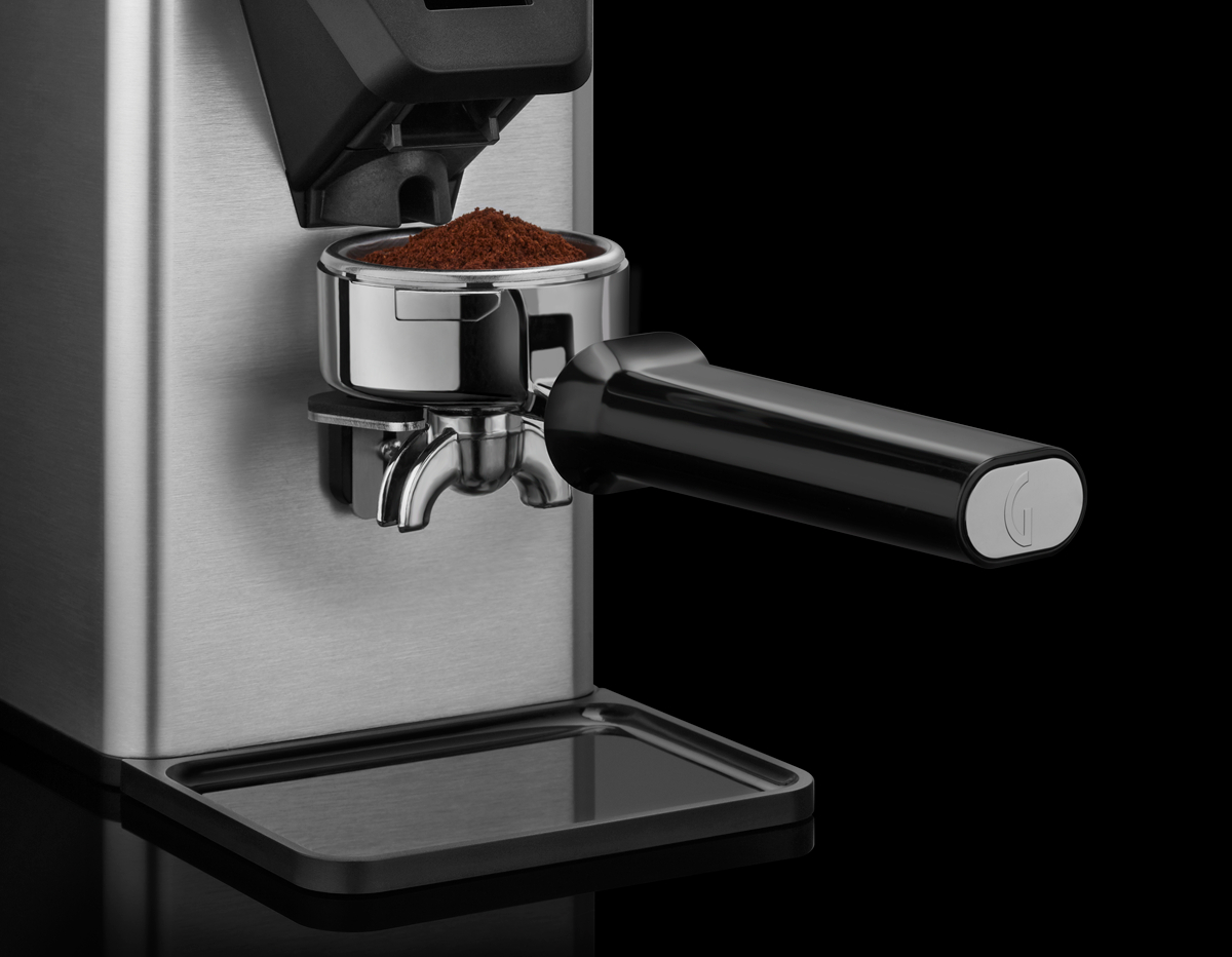 Eine Kaffeemühle aus Edelstahl mit einem Siebträger, der Kaffeepulver enthält. Ein schwarzer Griff erstreckt sich vom Siebträger.