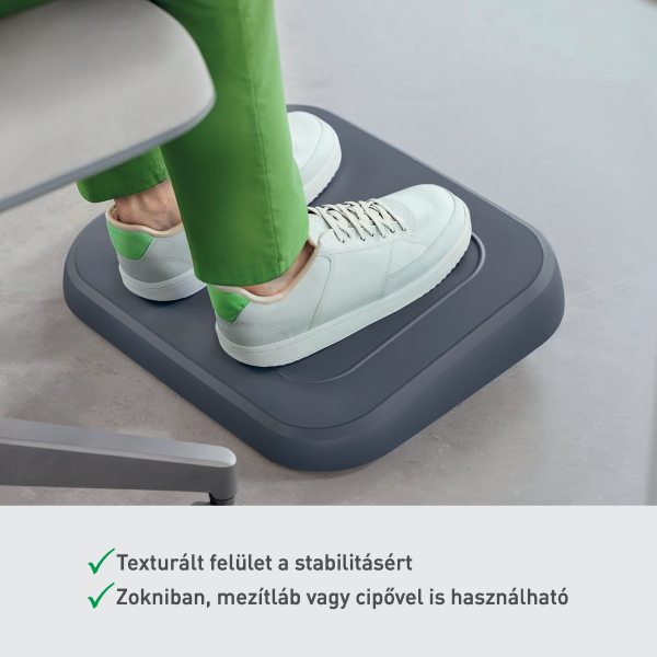 Fehér sportcipős személy lábai szürke lábtámaszon. Zöld nadrág látható. Szürke szék a háttérben, szürke padlón.