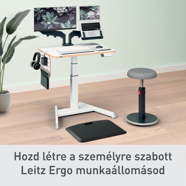 Fehér álló asztal két monitorral, laptop, fejhallgató és szék fa padlón. Egy cserepes növény balra.